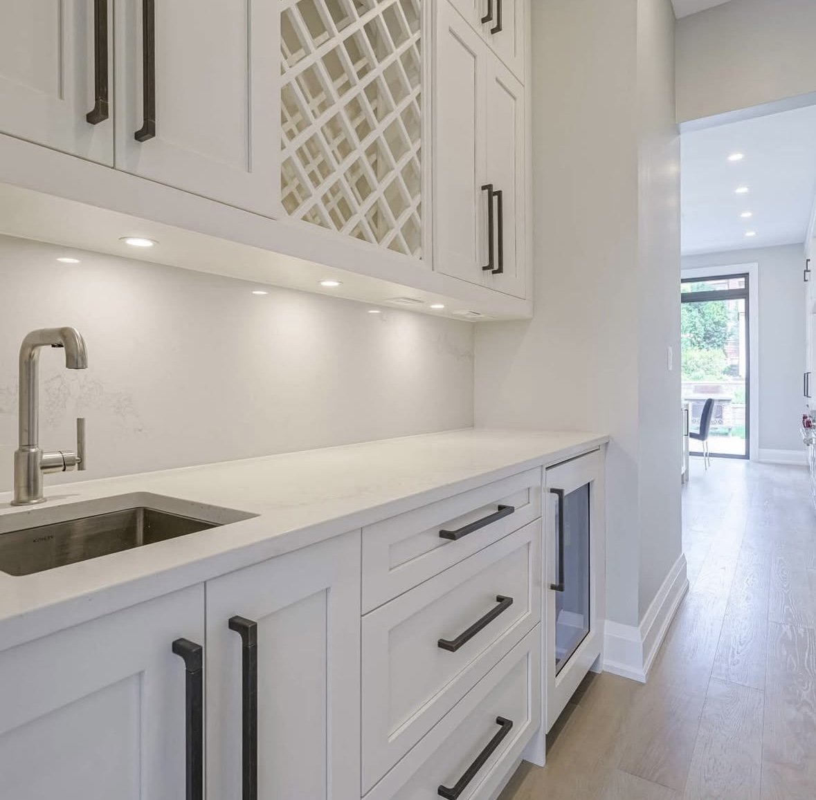 Kitchen pantry with white countertop, black handles, small sink, upper cabinets with integrated lighting and a view into a bright dining area with large window and sliding door.