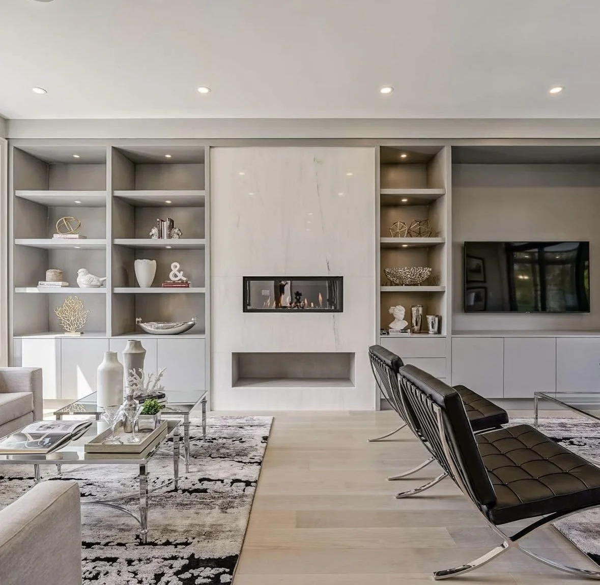 Modern amenity space /living room with a built-in white shelving unit, pot lights, fireplace, TV, black leather chairs, a beige sofa, a glass coffee table, and decorative objects on shelves.