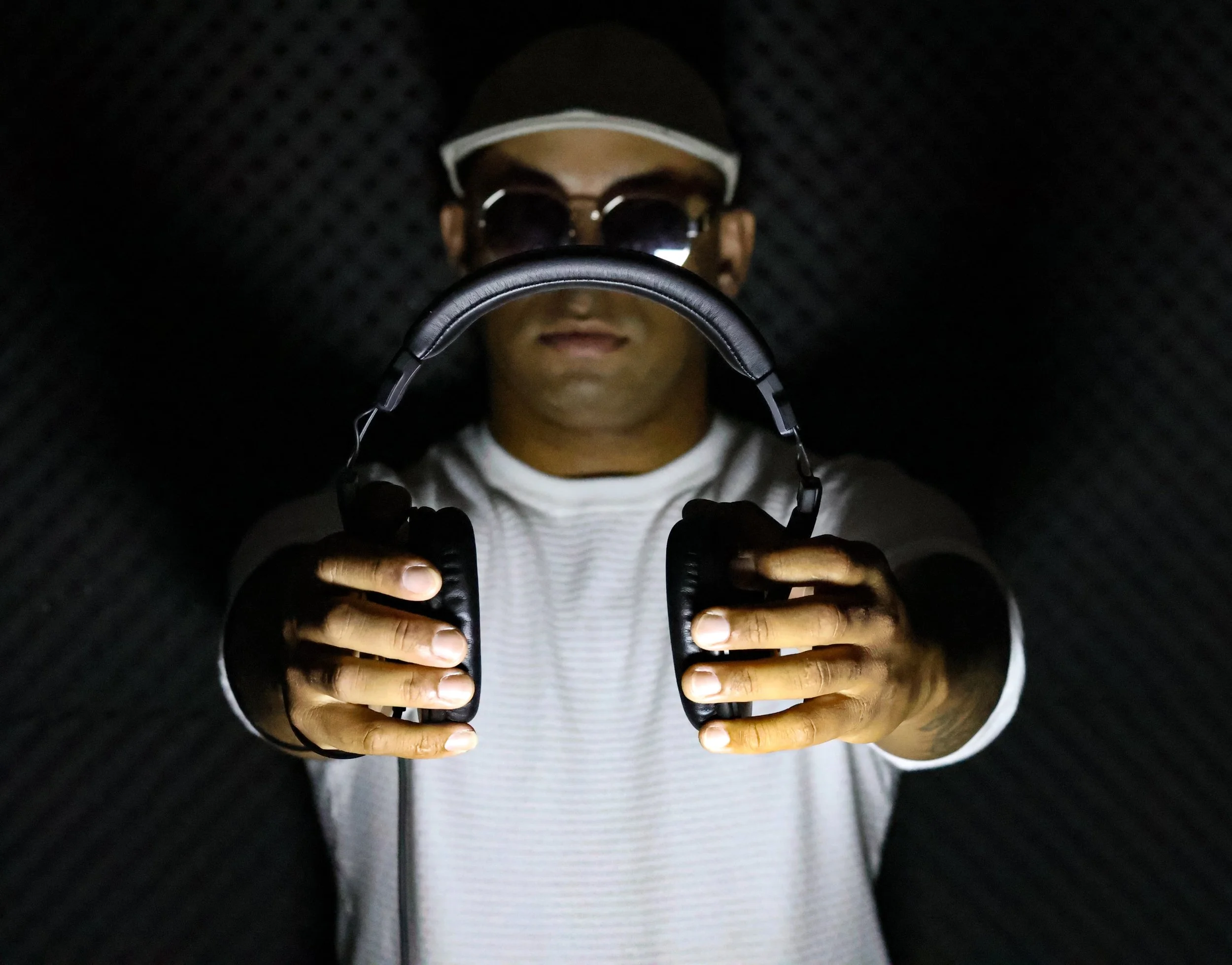 Pessoa com chapéu, óculos escuros, camiseta branca e cabelo agarrado, segurando fones de ouvido, em ambiente com paredes acústicas pretas.