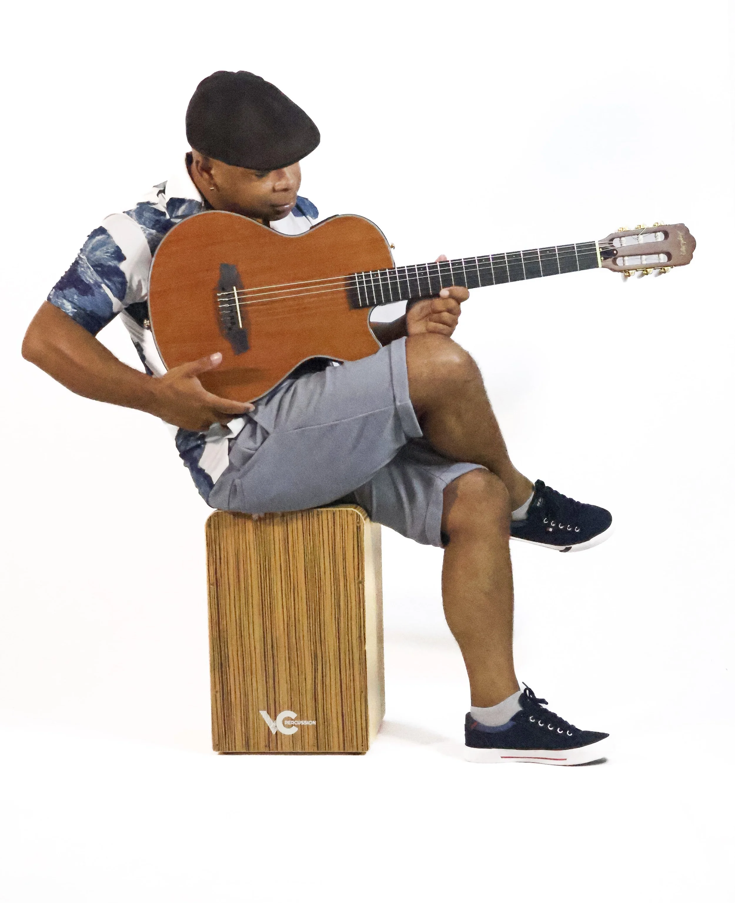 Pessoa tocando violão enquanto senta com uma perna cruzada, usando boné, camiseta de manga curta, bermuda cinza e tênis, sentado em um banquinho de madeira contra fundo branco.