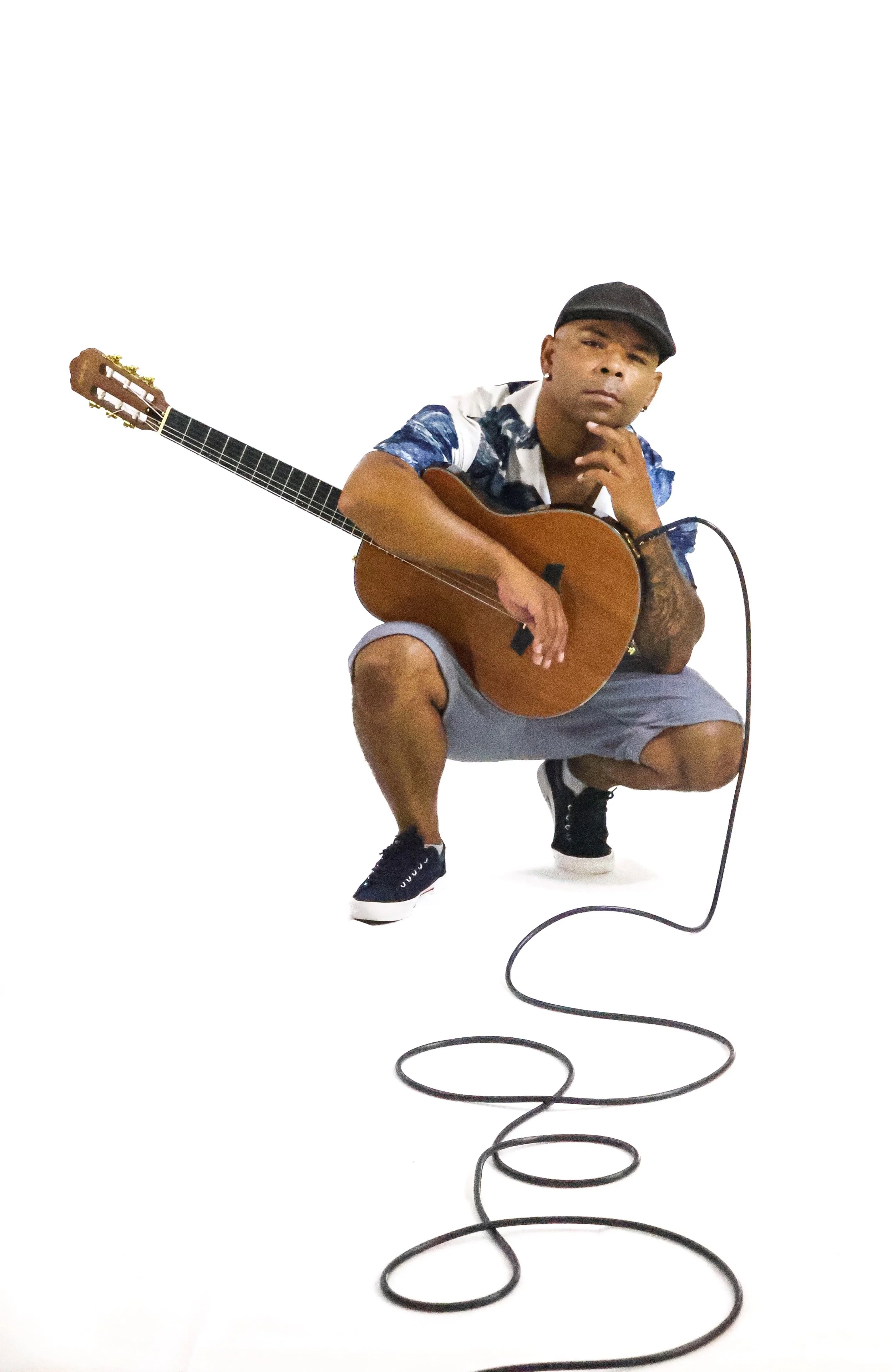 Homem com boné, camisa de seda azul e cinza, calça curta, e tênis pretos, segurando uma guitarra acústica, em pose com expressão pensativa, com um microfone no chão e fundo branco