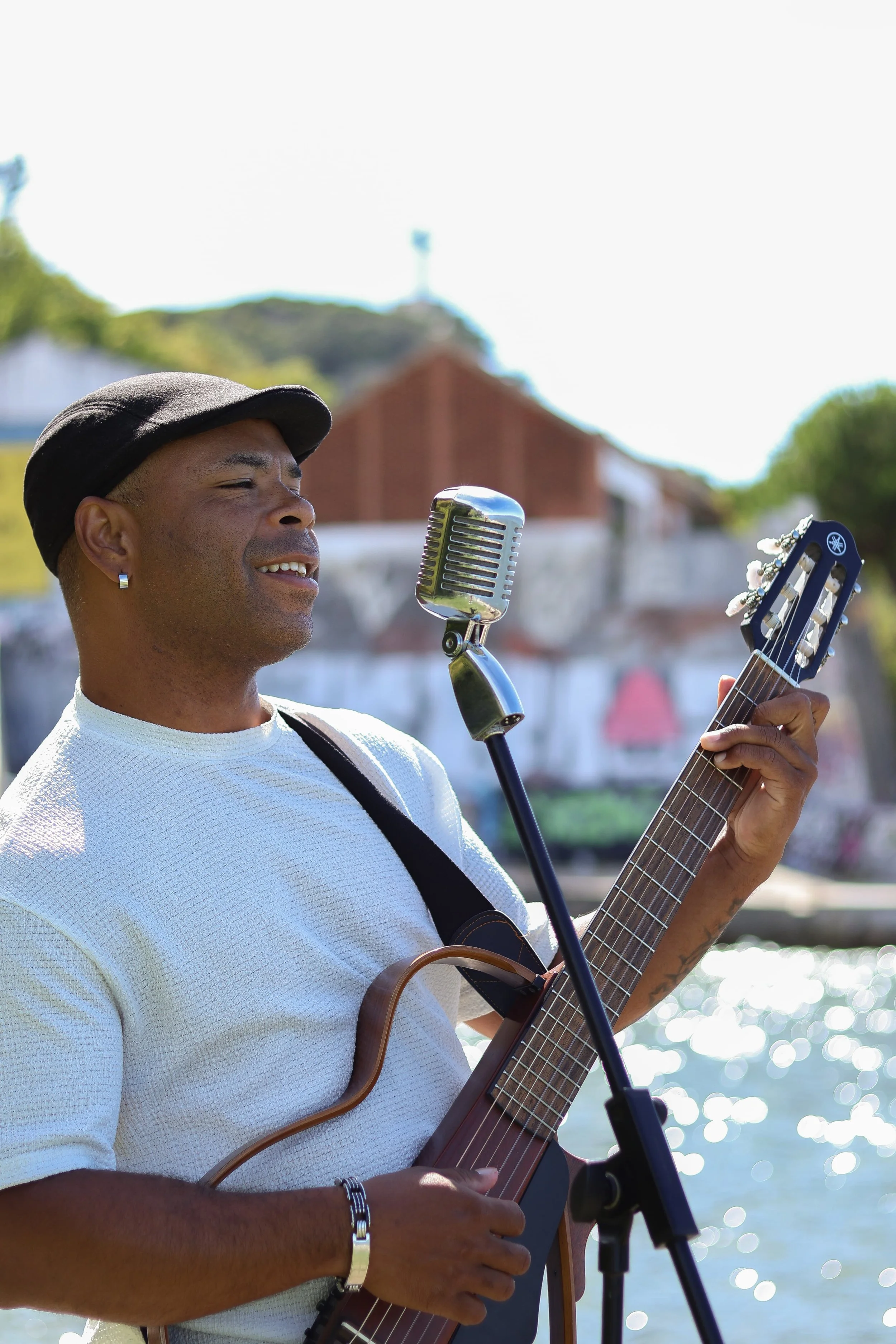 Homem tocando violão e cantando em frente a uma microfone ao ar livre, com rio e casas ao fundo.