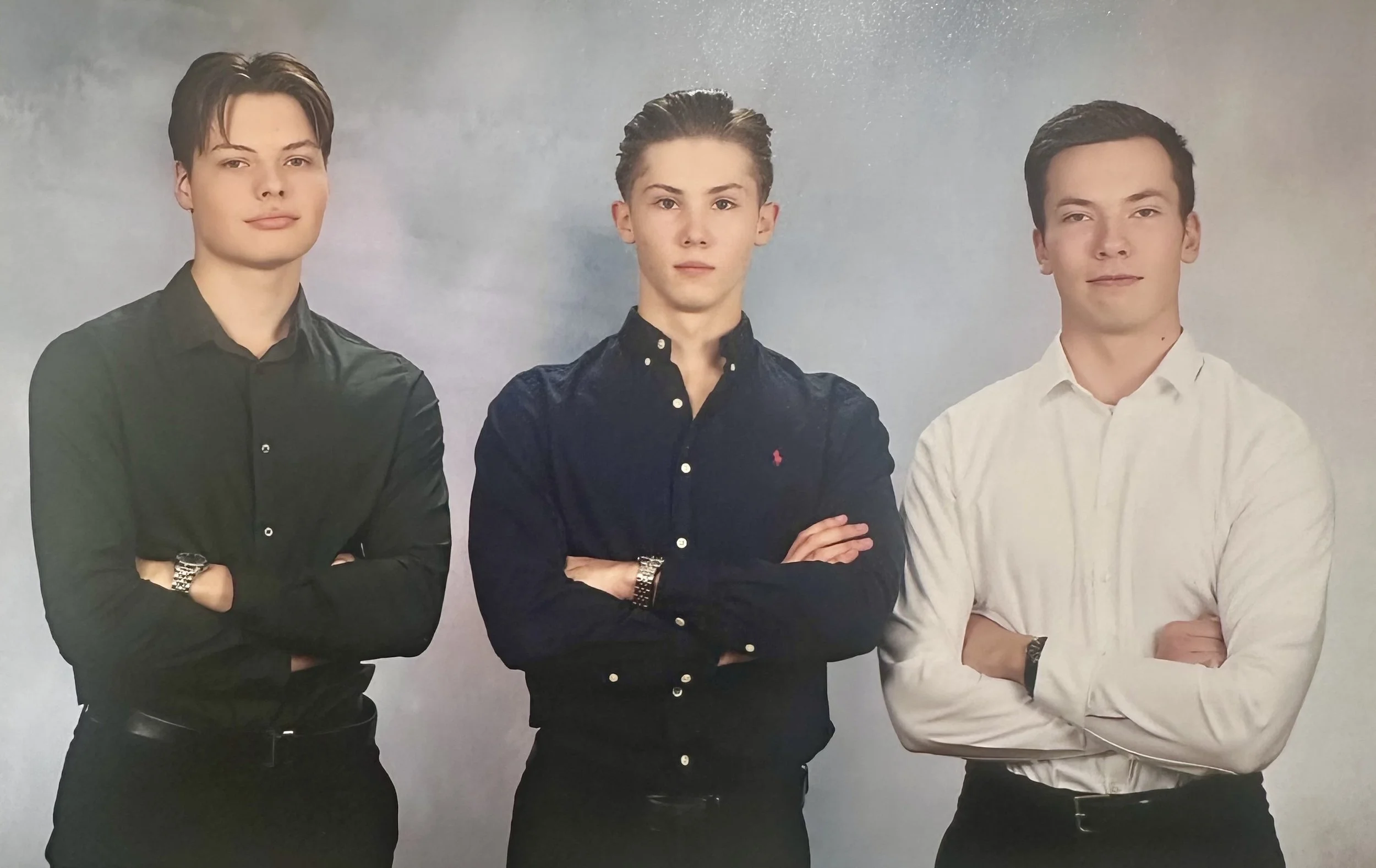 Three young men standing with arms crossed, dressed in formal shirts, against a plain gray background.