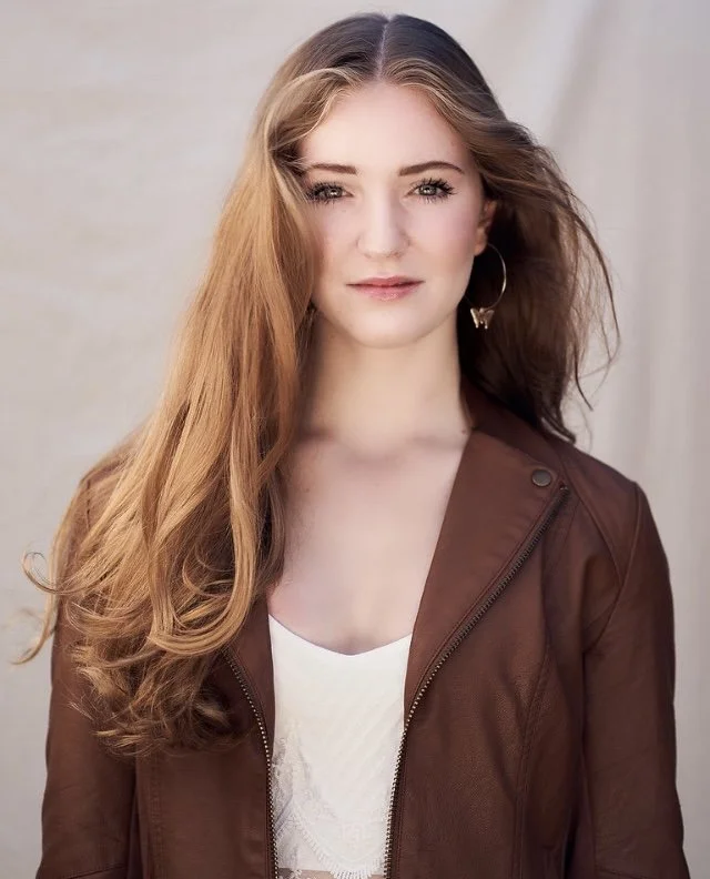 A young woman with long, wavy red hair wearing a brown jacket over a white top, standing against a neutral background.