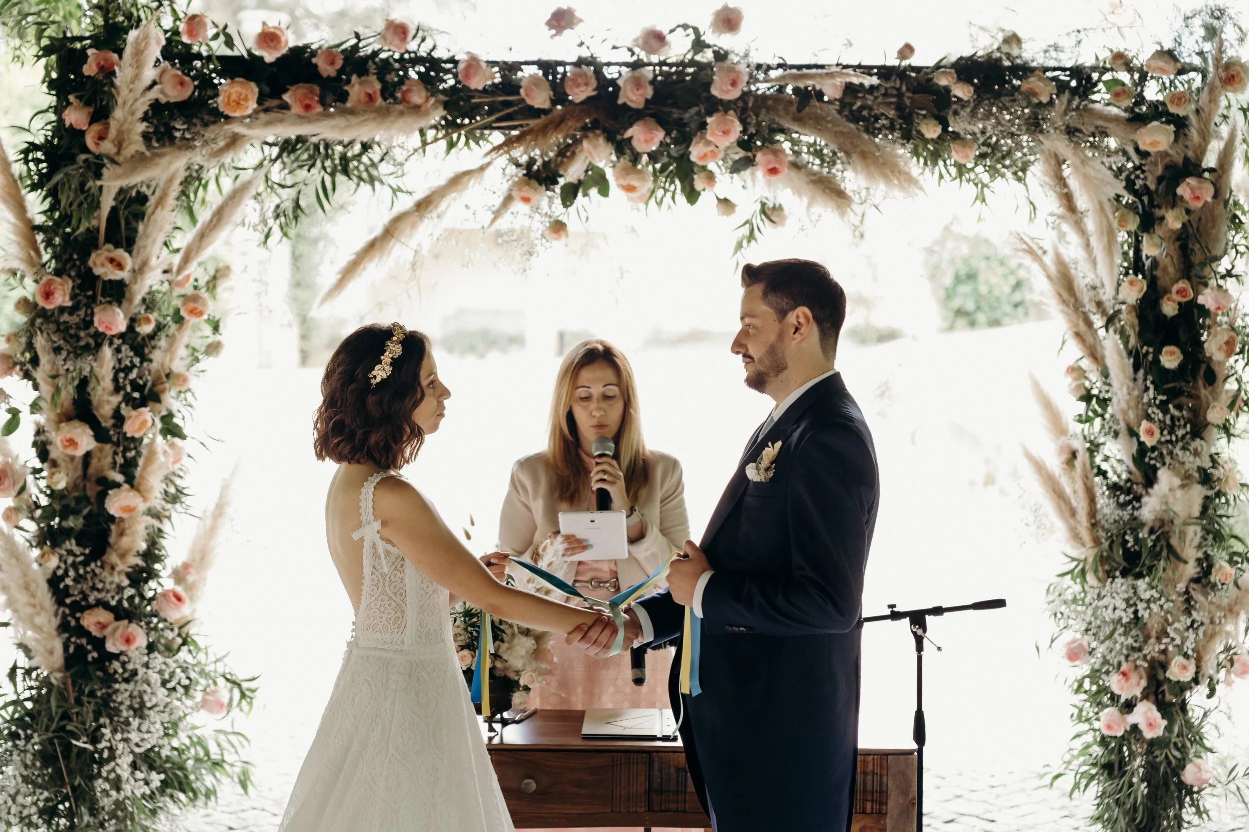A bride and groom holding hands during their wedding ceremony, standing under a floral arch with an officiant reading from a tablet, outdoors in bright daylight.