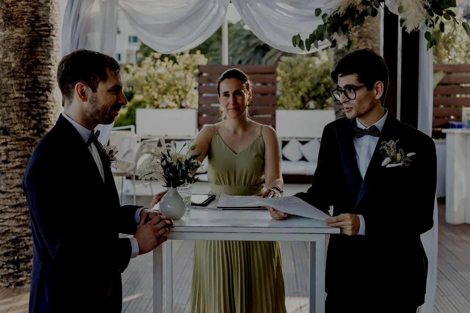 Two men in tuxedos holding hands during an outdoor wedding ceremony, with a woman officiant standing behind them, under a decorated canopy, with trees and outdoor furniture in the background.