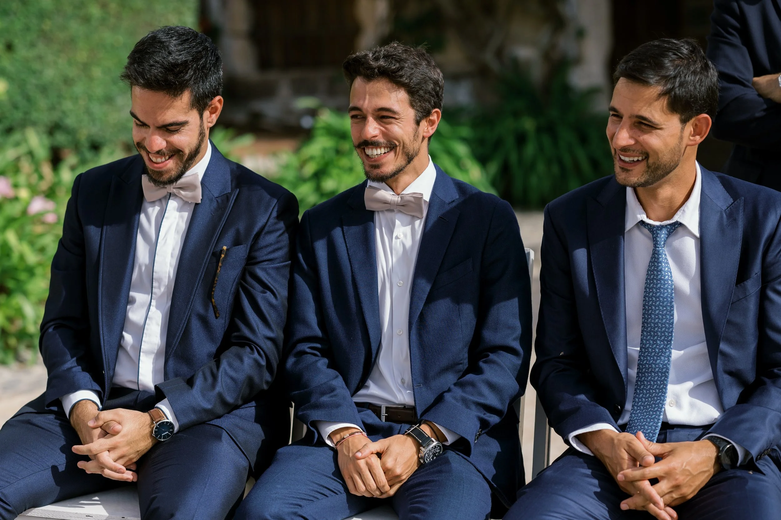 Three men in suits and bow ties sitting outdoors, smiling and looking down.