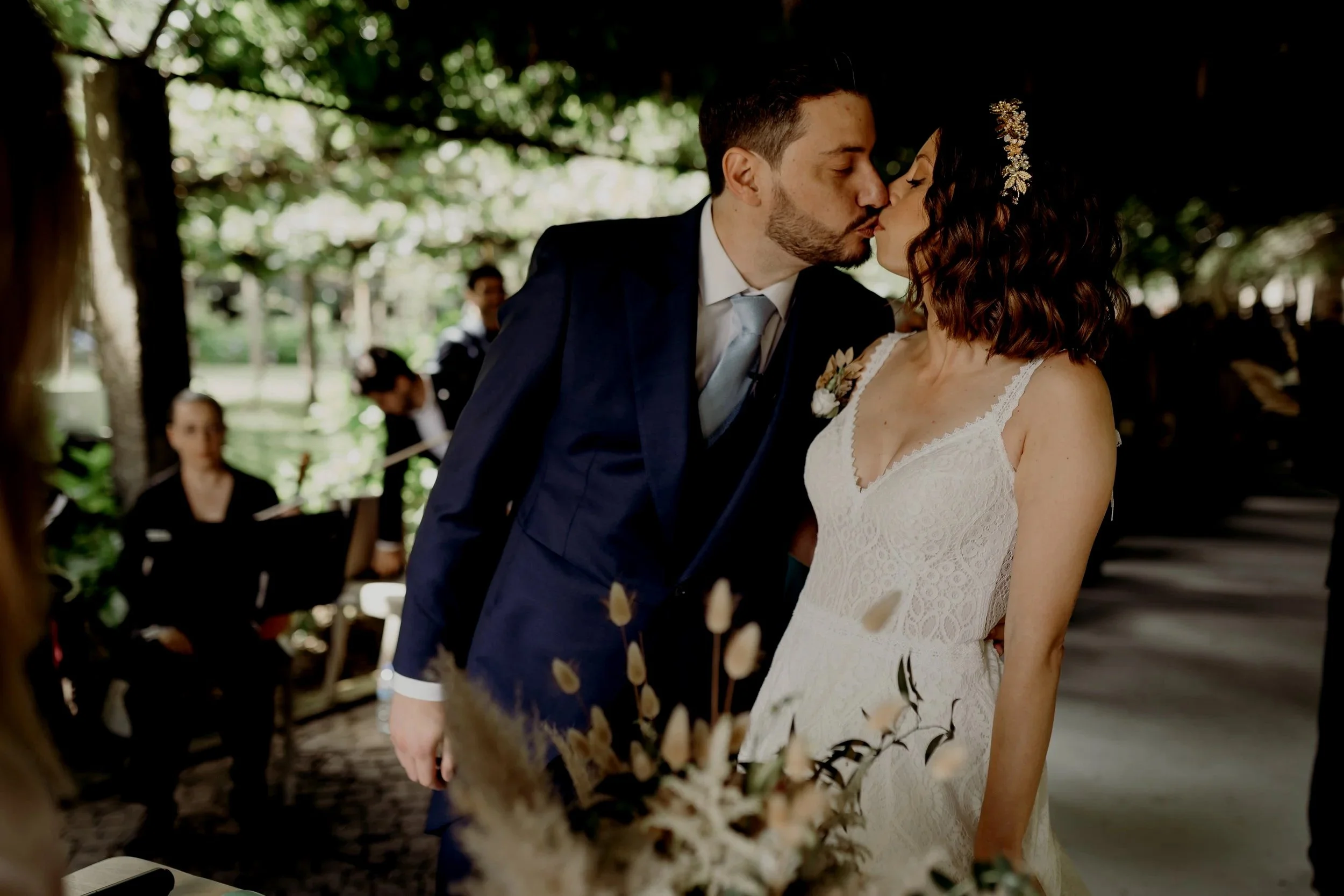 A couple shares a kiss during a wedding ceremony outdoors, with guests and musicians in the background.