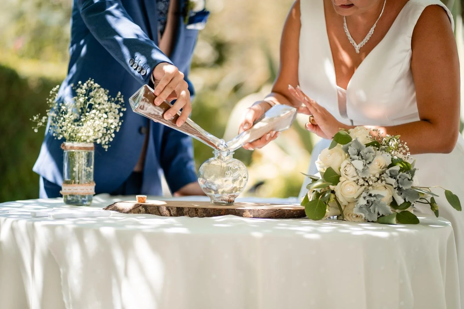 A wedding ceremony with a bride and groom pouring sand into a glass vase as part of a unity sand ritual, with floral decorations on a table outdoors.