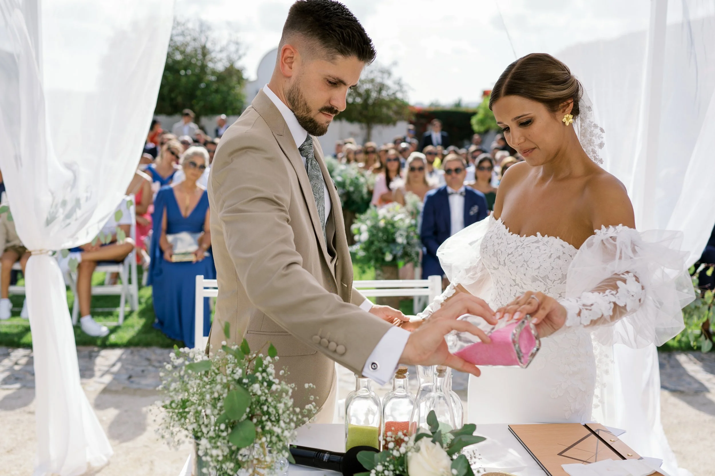A bride and groom exchanging rings during an outdoor wedding ceremony, with guests seated in the background.