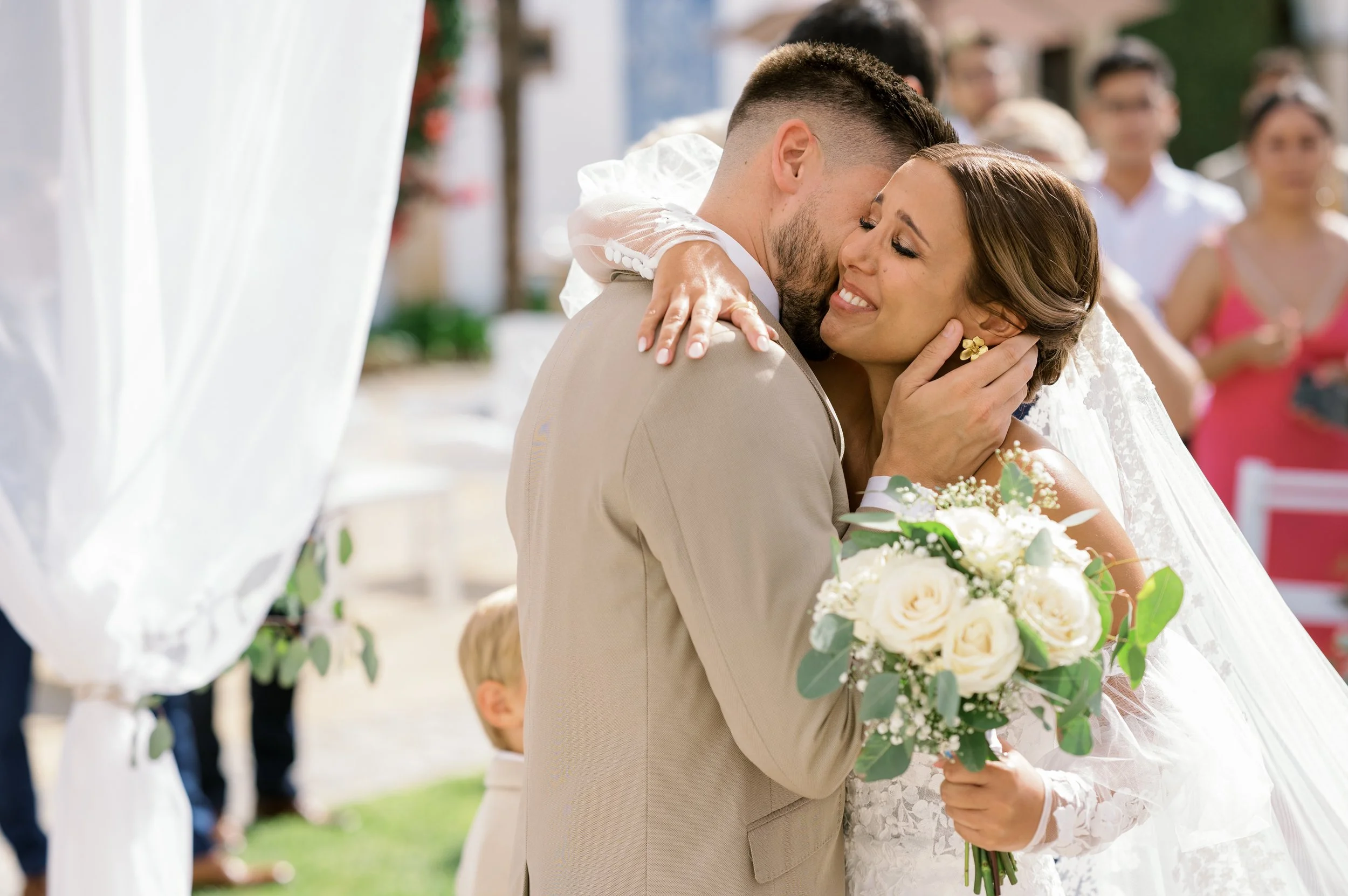 A bride and groom sharing a moment during their wedding, with the bride holding a bouquet of white roses and wearing a lace dress, while the groom wears a beige suit, outdoors with guests in the background.