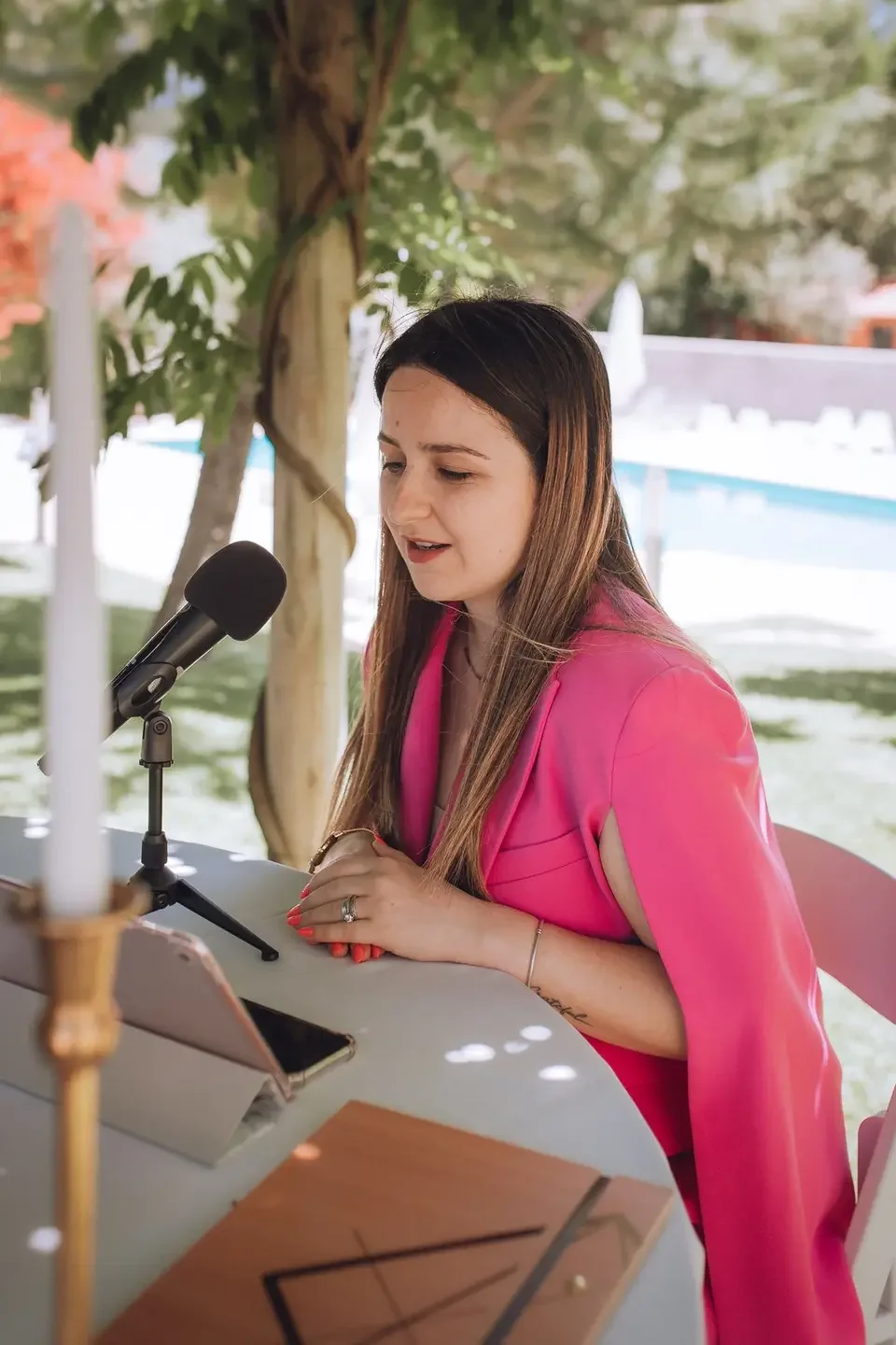 Young woman with long brown hair and a pink dress speaking into a microphone during an outdoor event
