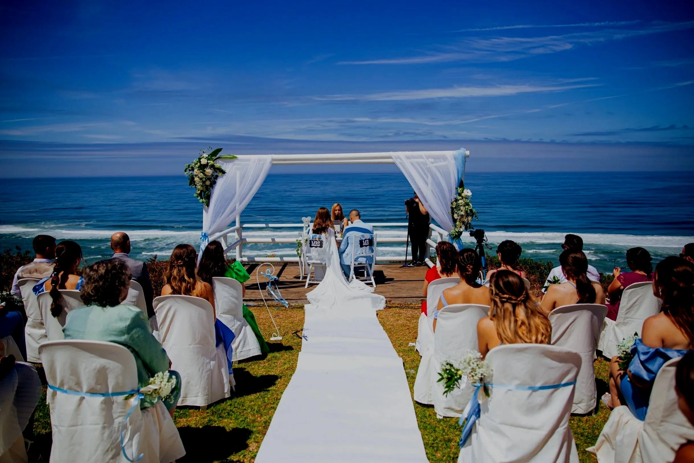 A couple kisses at their wedding ceremony outdoors, with wedding officiants seated and greenery around.