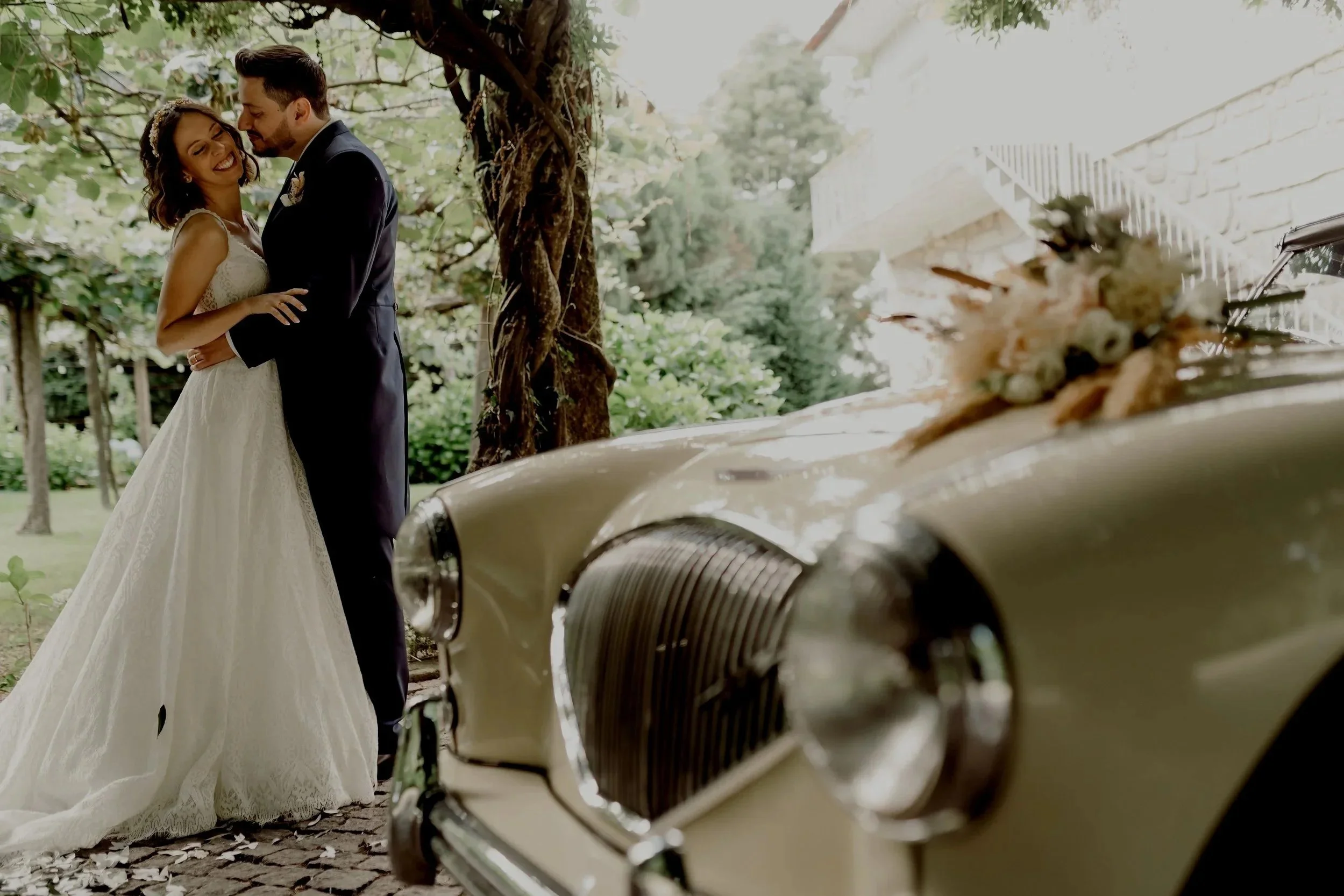 A bride and groom embracing outdoors near a vintage car decorated with flowers and greenery, with trees and a building in the background.