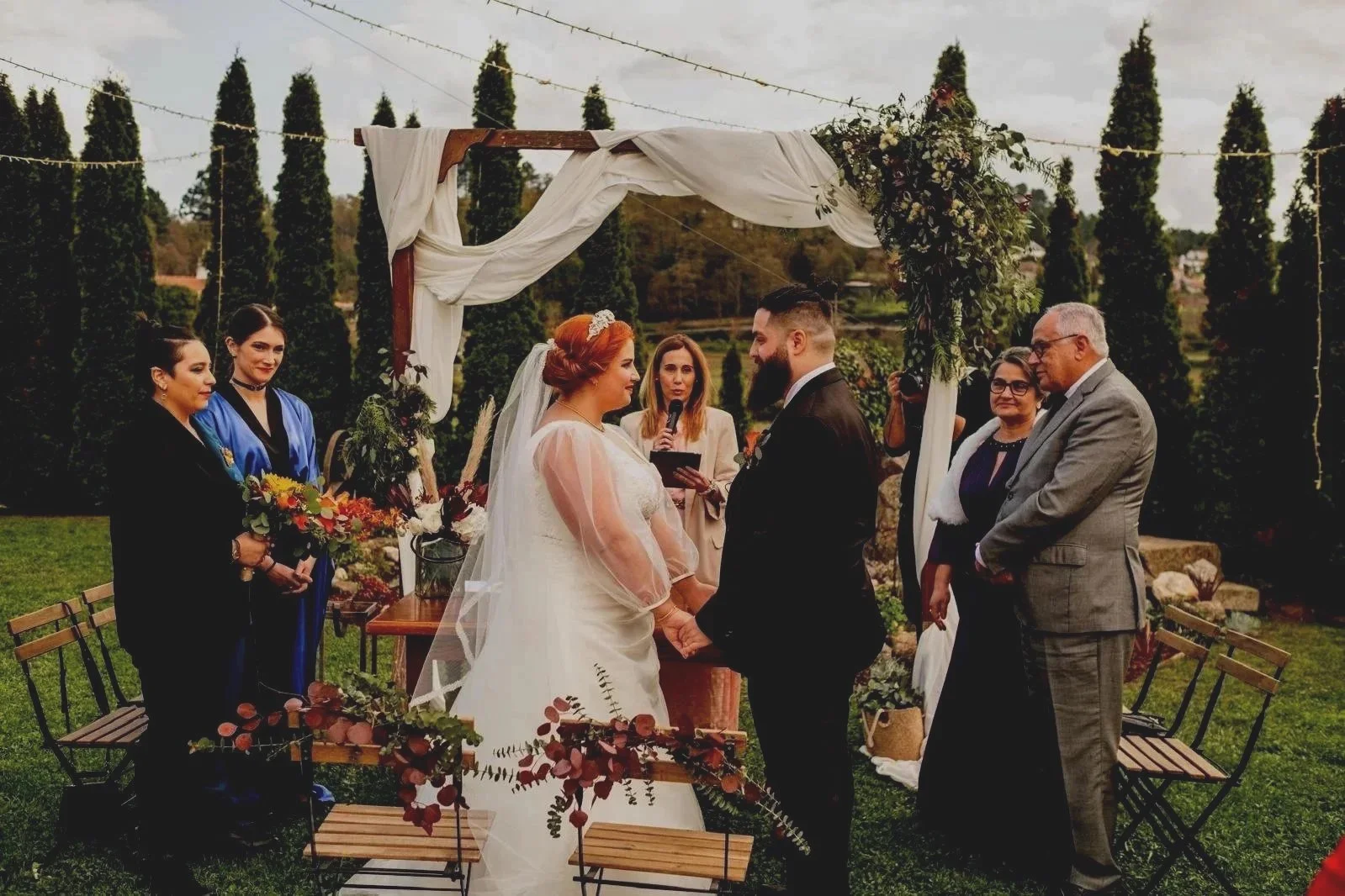 A couple getting married outdoors under a decorated arch, holding hands and gazing at each other, with a woman officiating in the background. Guests and wedding party members stand nearby, with chairs and floral arrangements around, against a backdrop of tall trees and a cloudy sky.
