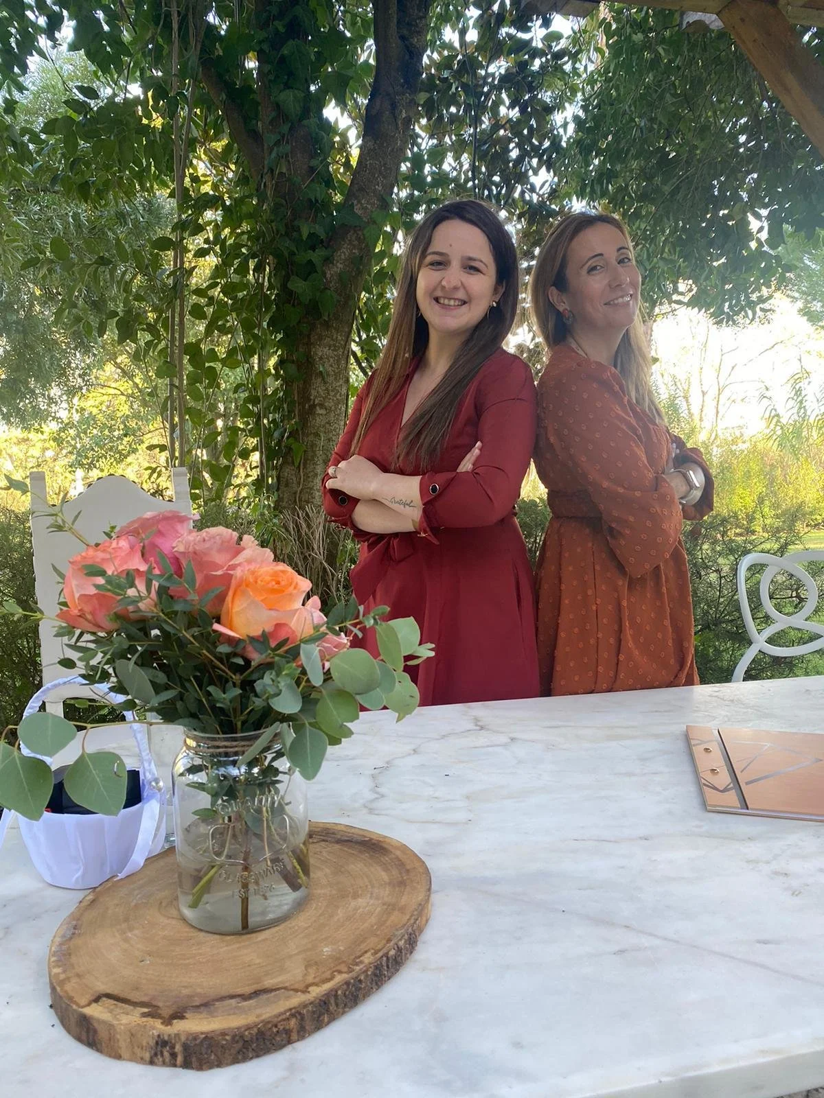 Two women standing outdoors at a table, back-to-back, smiling at the camera. One woman has dark hair and is wearing a red dress; the other has light hair and is wearing a brown dress with a pattern. On the white table in front of them is a glass jar with a bouquet of pink and peach roses and greenery, placed on a round wooden slab.