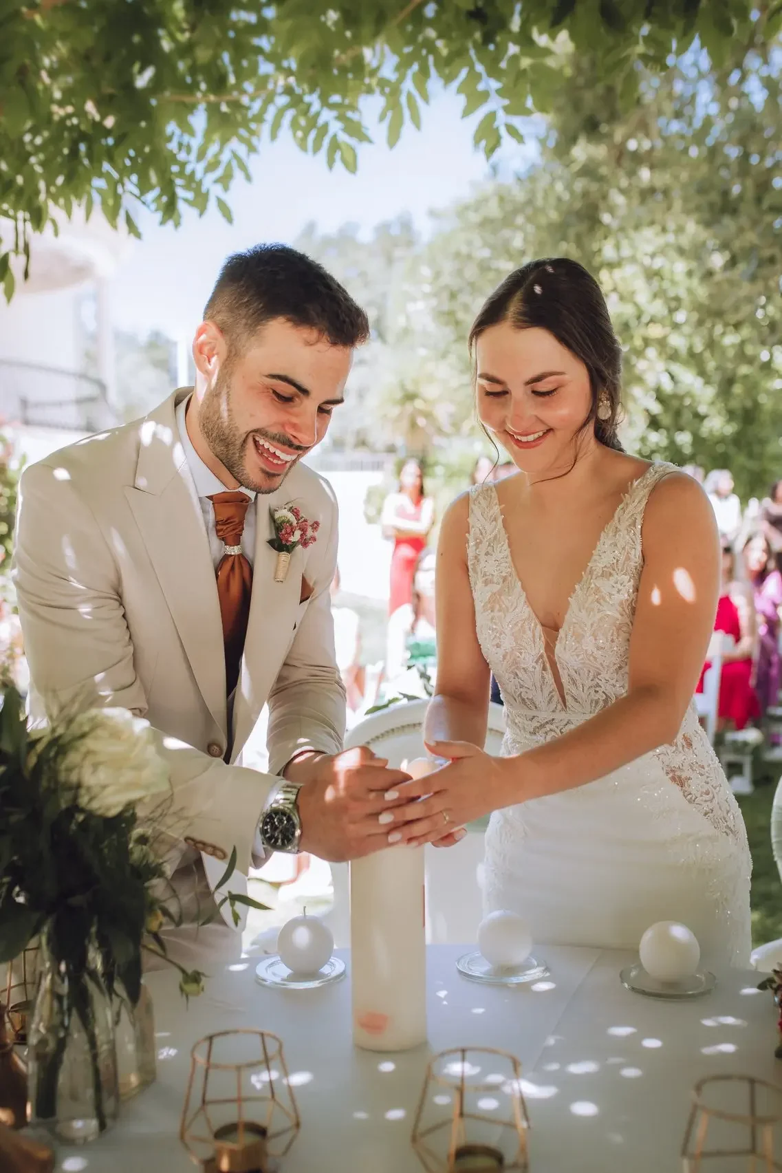A bride and groom are smiling and holding hands during their wedding ceremony outdoors, with trees and guests in the background.