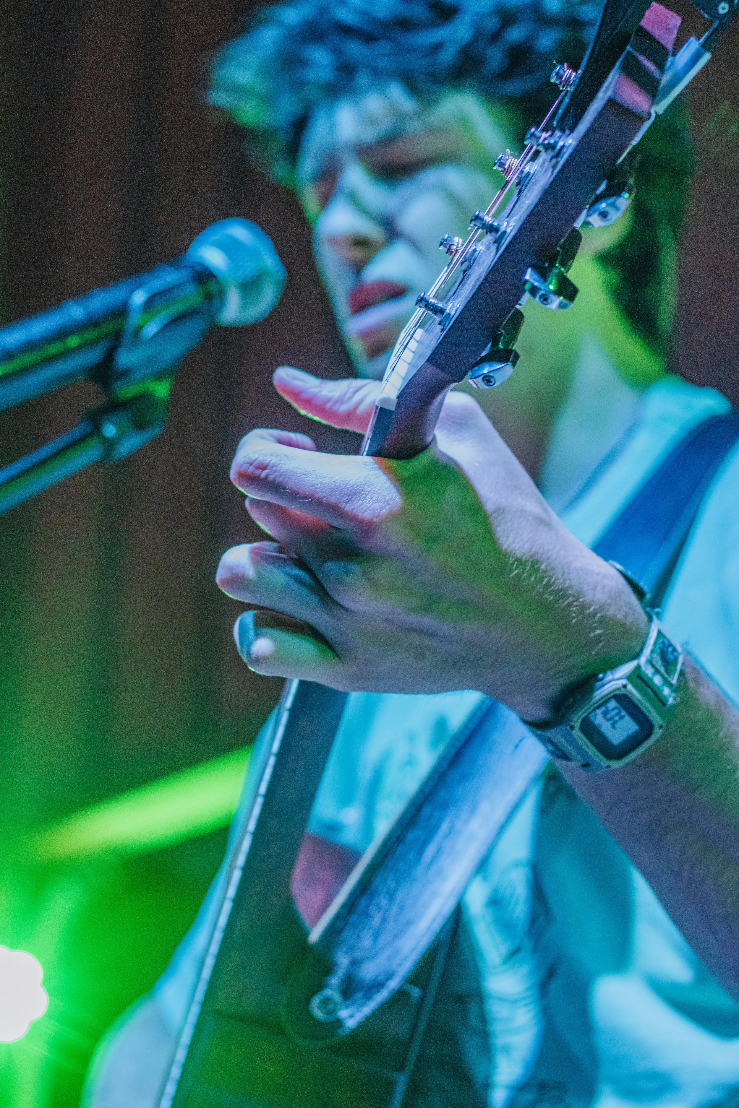 A close-up of a person playing an acoustic guitar on stage, with a microphone in front and colorful stage lights illuminating the scene.