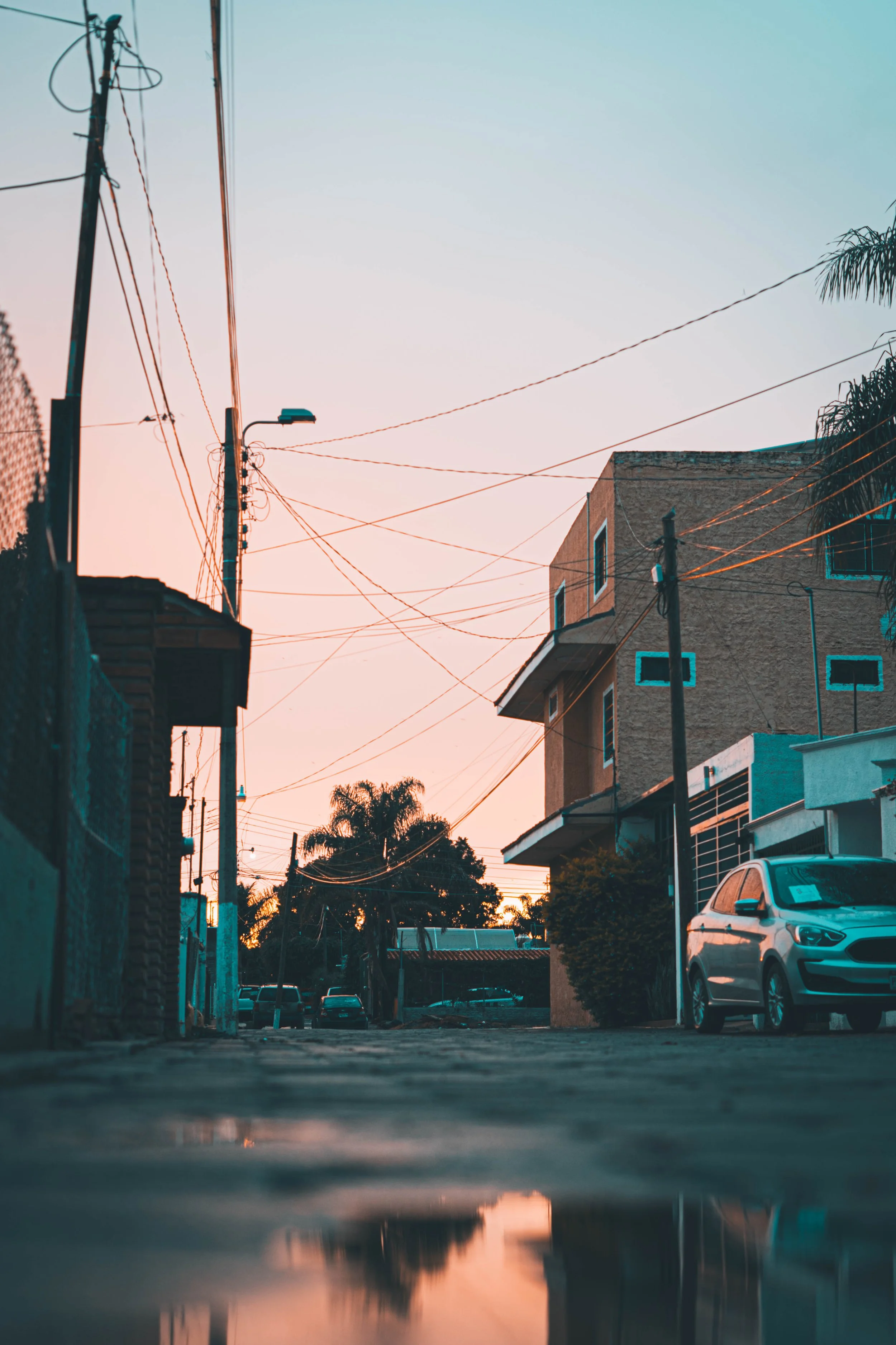 A quiet urban street scene at sunset with power lines, buildings, trees, and a parked car.