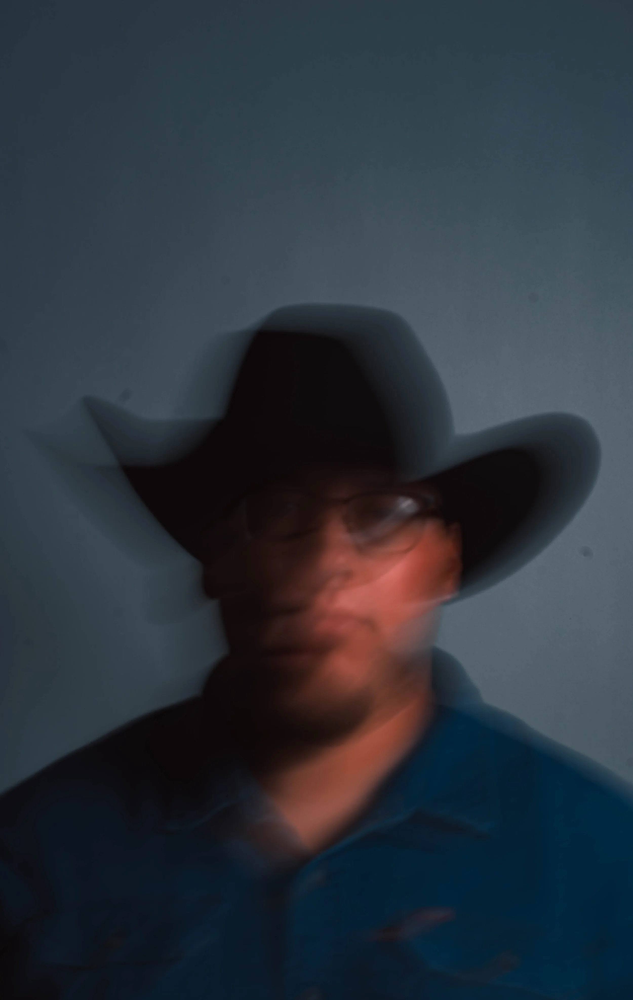 Blurry photo of a person wearing a wide-brimmed hat and glasses, with a dark background.