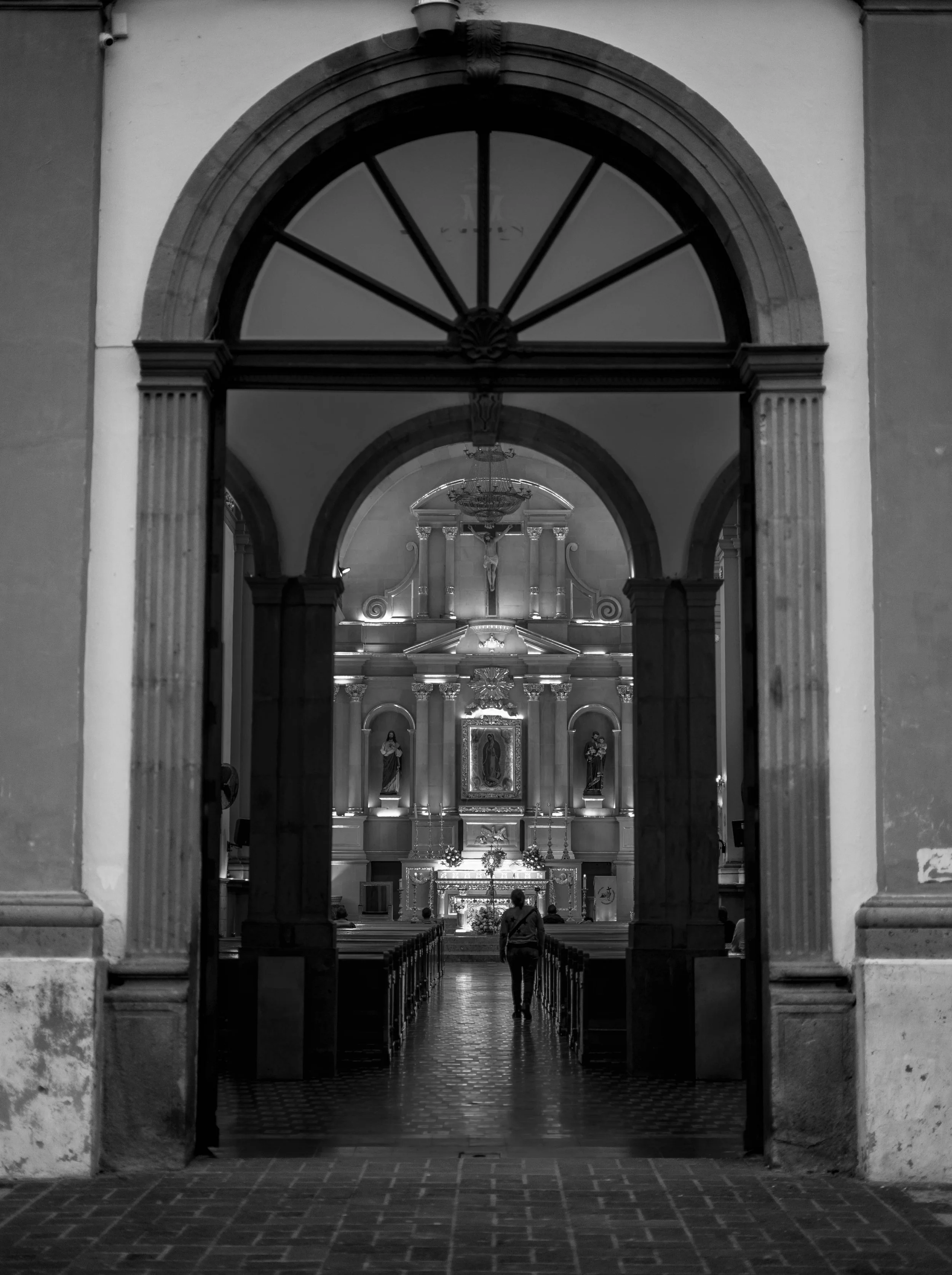 Inside view of a church through an arched doorway, showing a person walking toward the altar with religious statues and a crucifix.