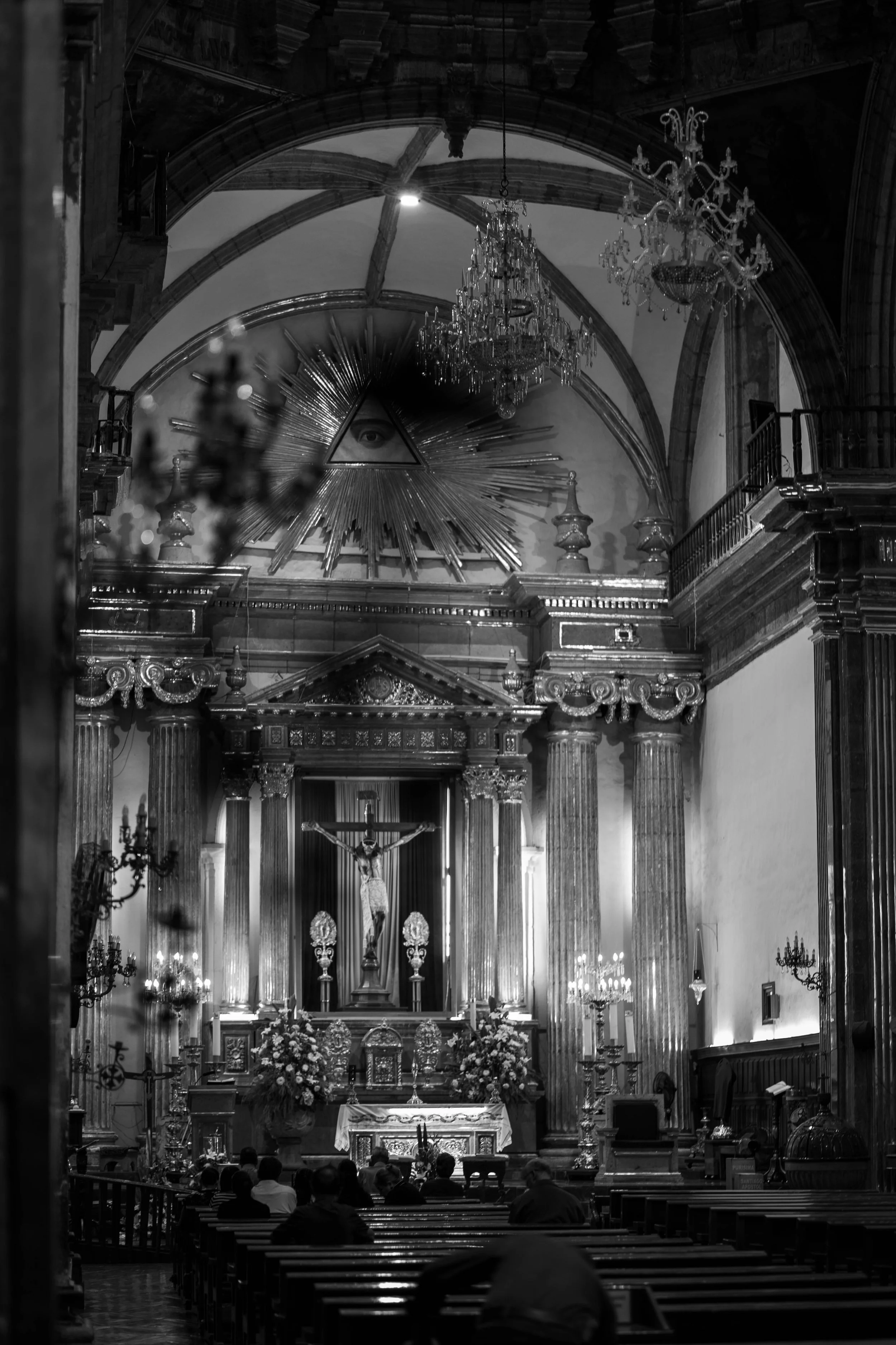 Interior of a church with altar, crucifix, columns, chandeliers, and religious symbols, depicted in black and white.