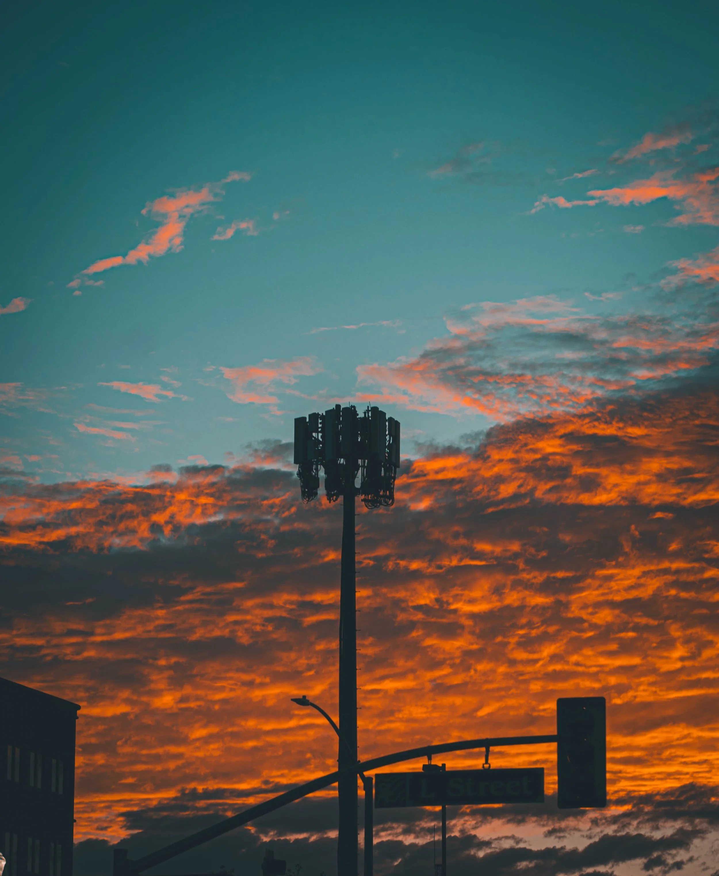 A sunset sky with orange and blue clouds, a tall cellular tower, a street sign, and a traffic light.