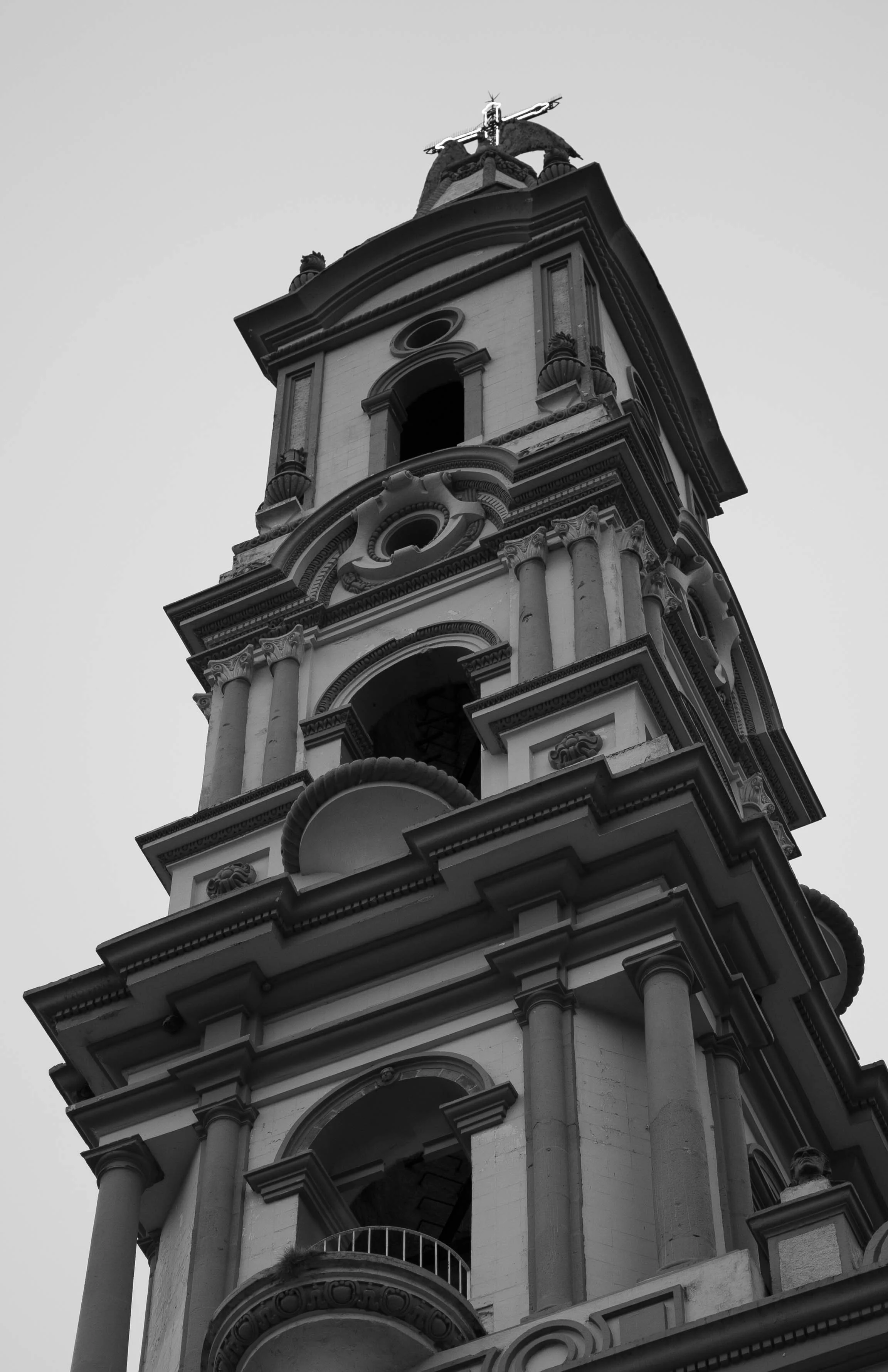 Black and white photo of a tall, ornate church tower with multiple levels, columns, decorative moldings, and a cross at the top.