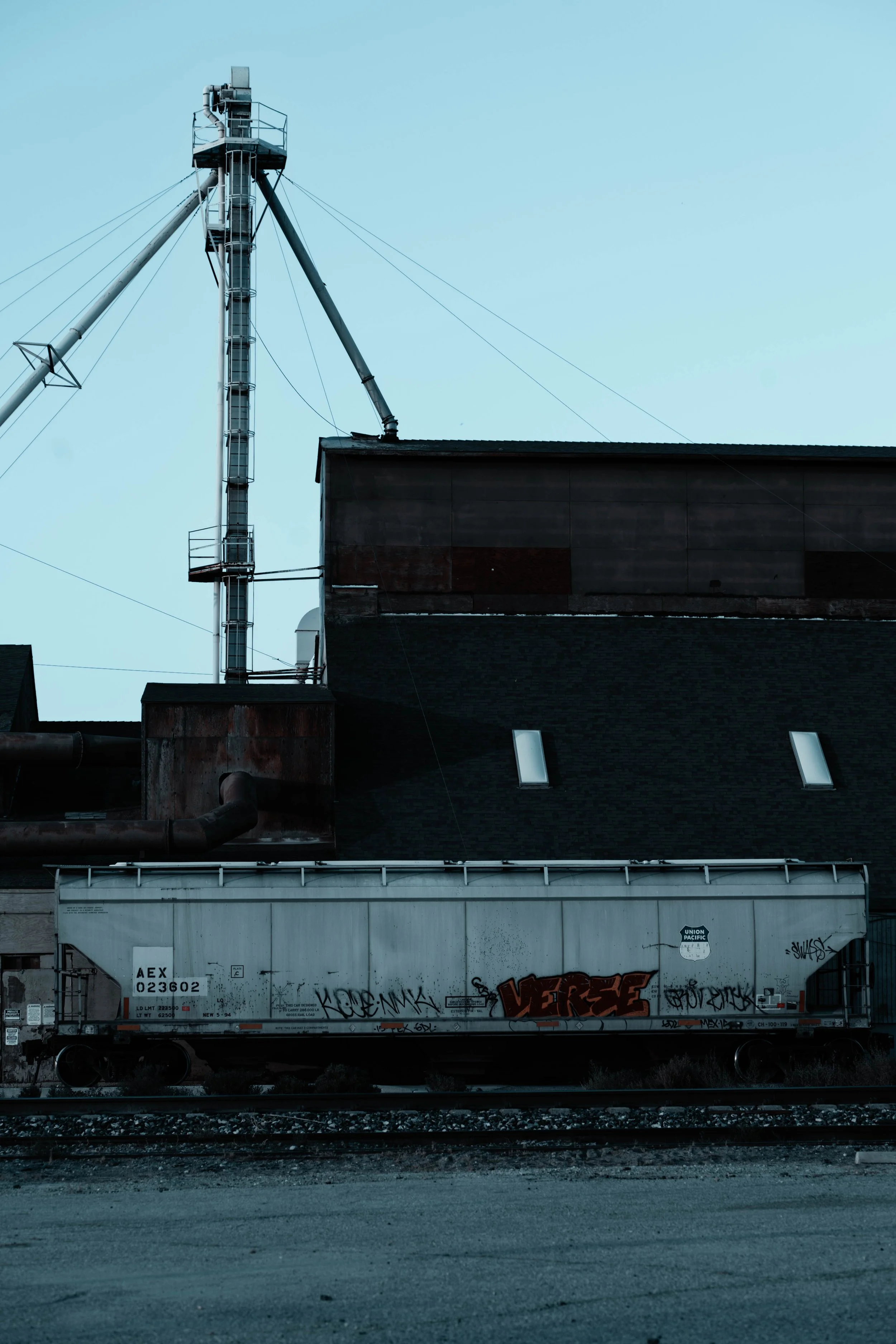 An industrial building with a rooftop crane, graffiti-covered train car in front, and railway tracks at the bottom against a clear sky.