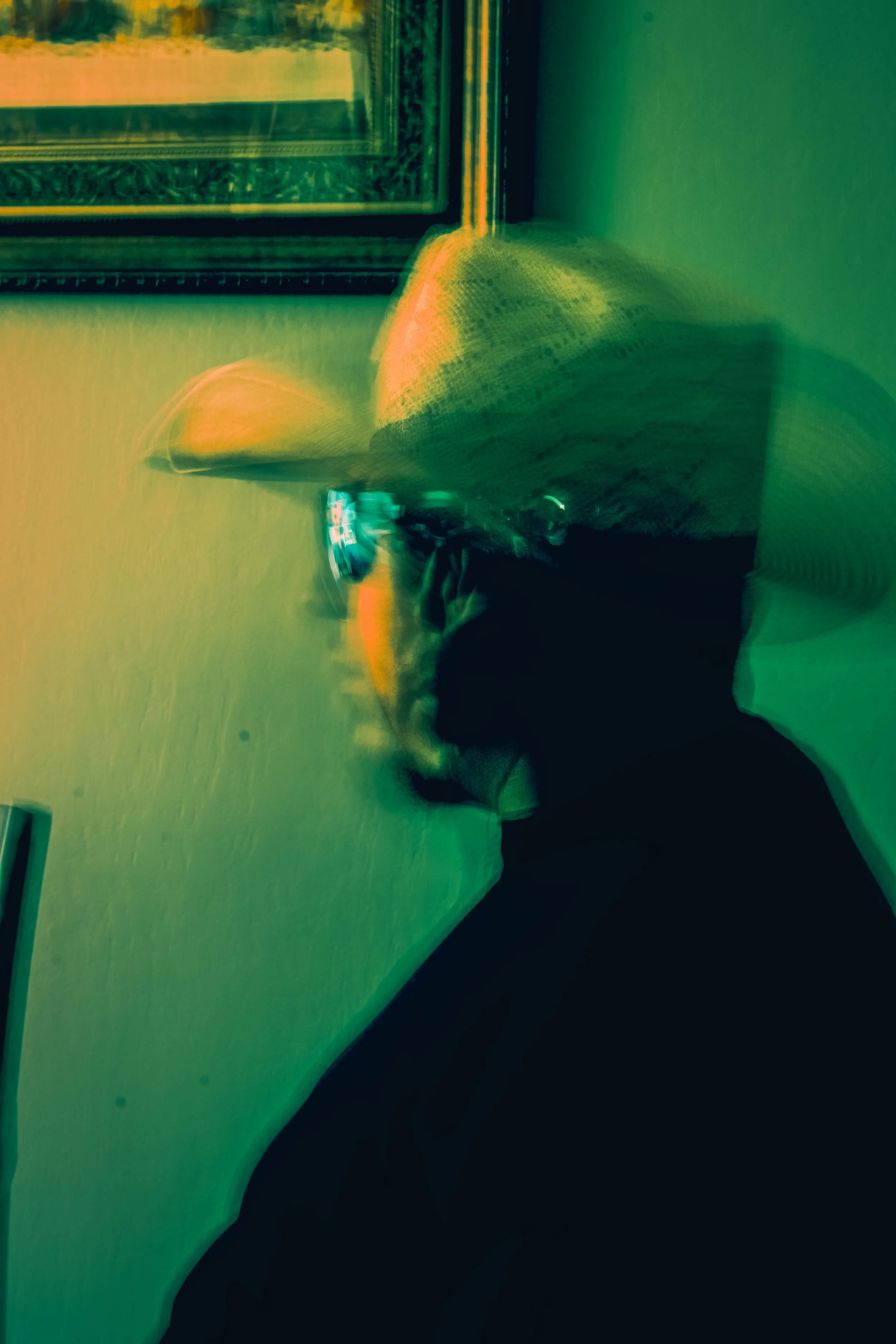 Person sitting on a couch wearing sunglasses, holding a smartphone, with a picture frame on the wall in the background, and the photo has a colorful, abstract, distorted effect.