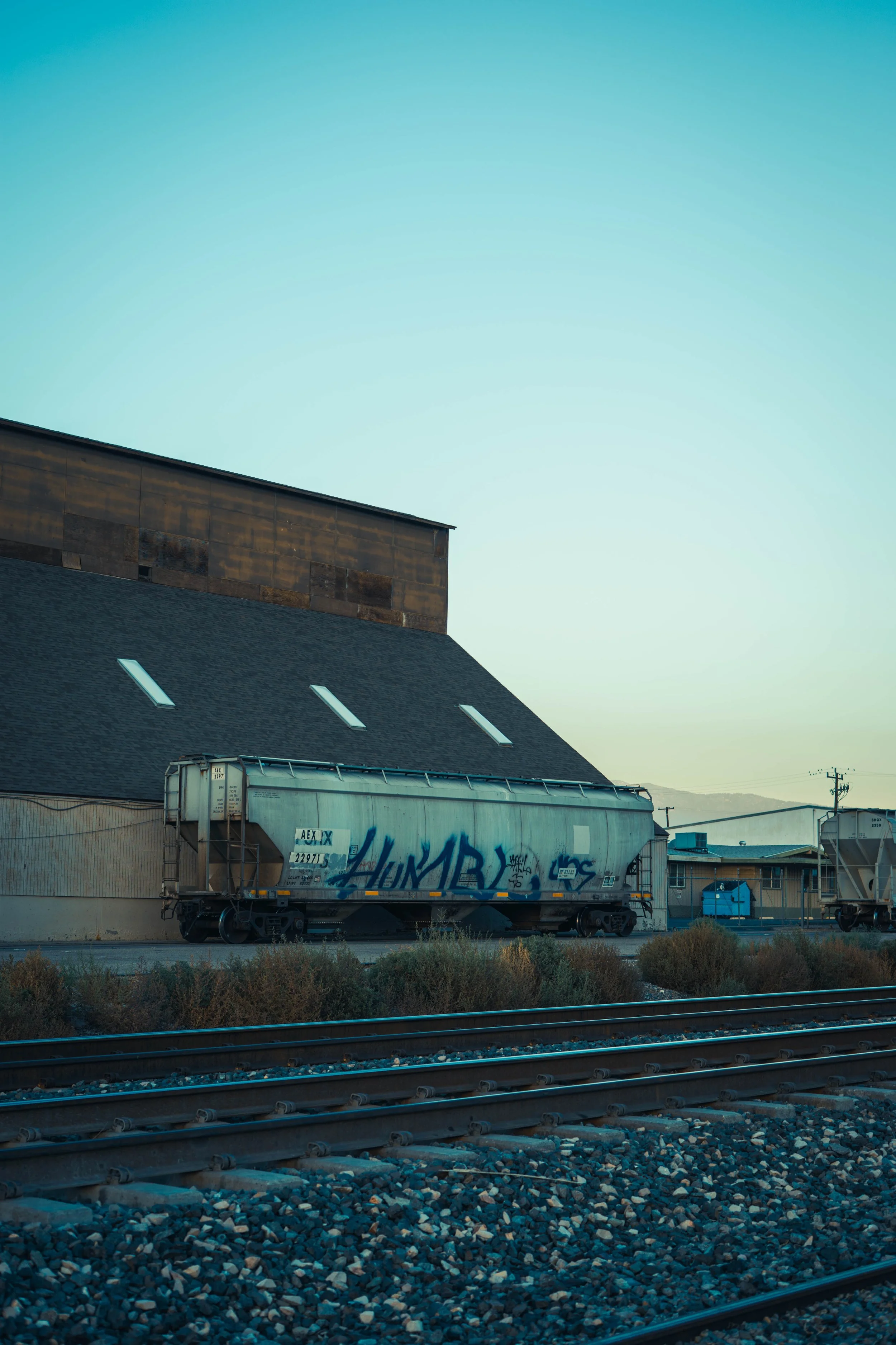 A graffitied train car parked on railroad tracks beside a building, with a blue sky overhead.