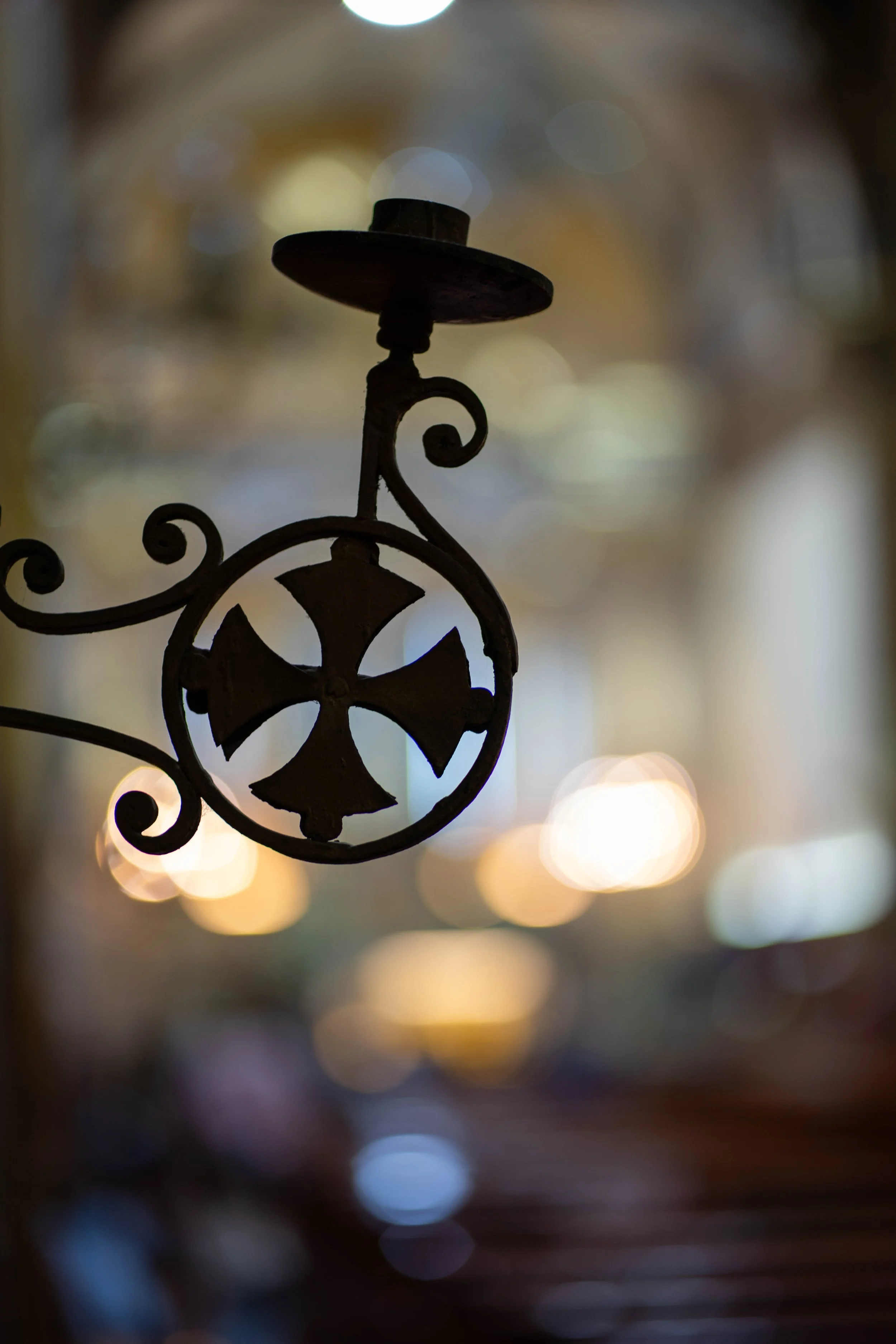 Close-up of a metal candle holder with a cross design, hanging in front of a blurred background with circular bokeh lights.