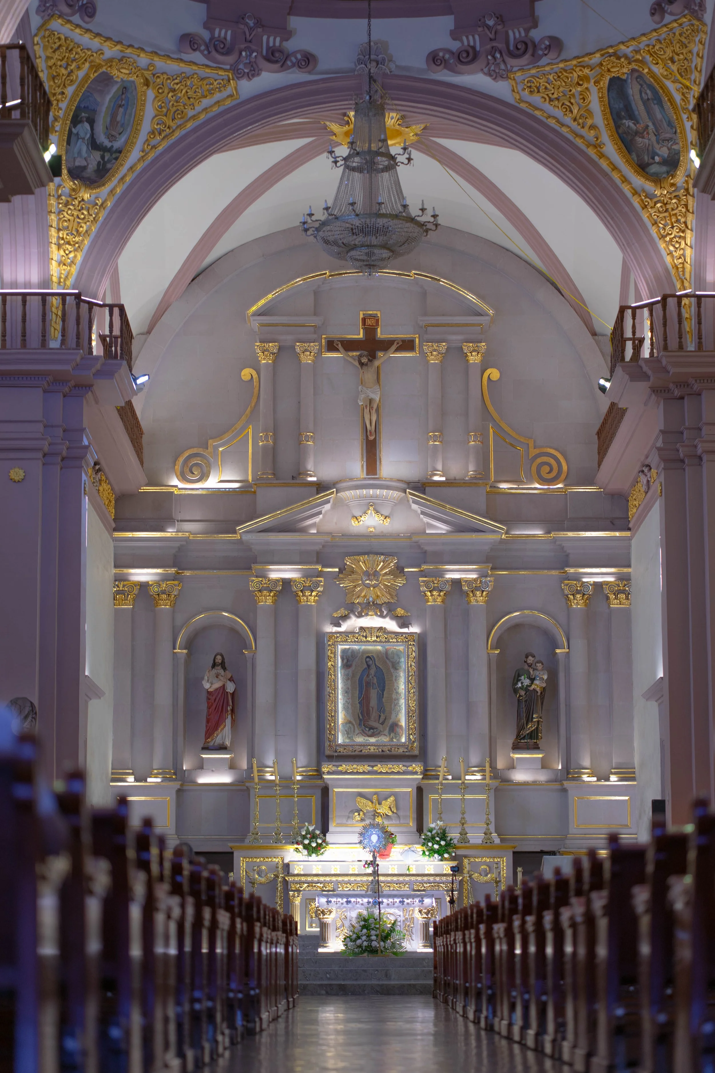 Interior of a Catholic church with an ornate altar featuring gold accents, statues of religious figures, a crucifix with Jesus on the cross, and a large chandelier hanging from the ceiling.