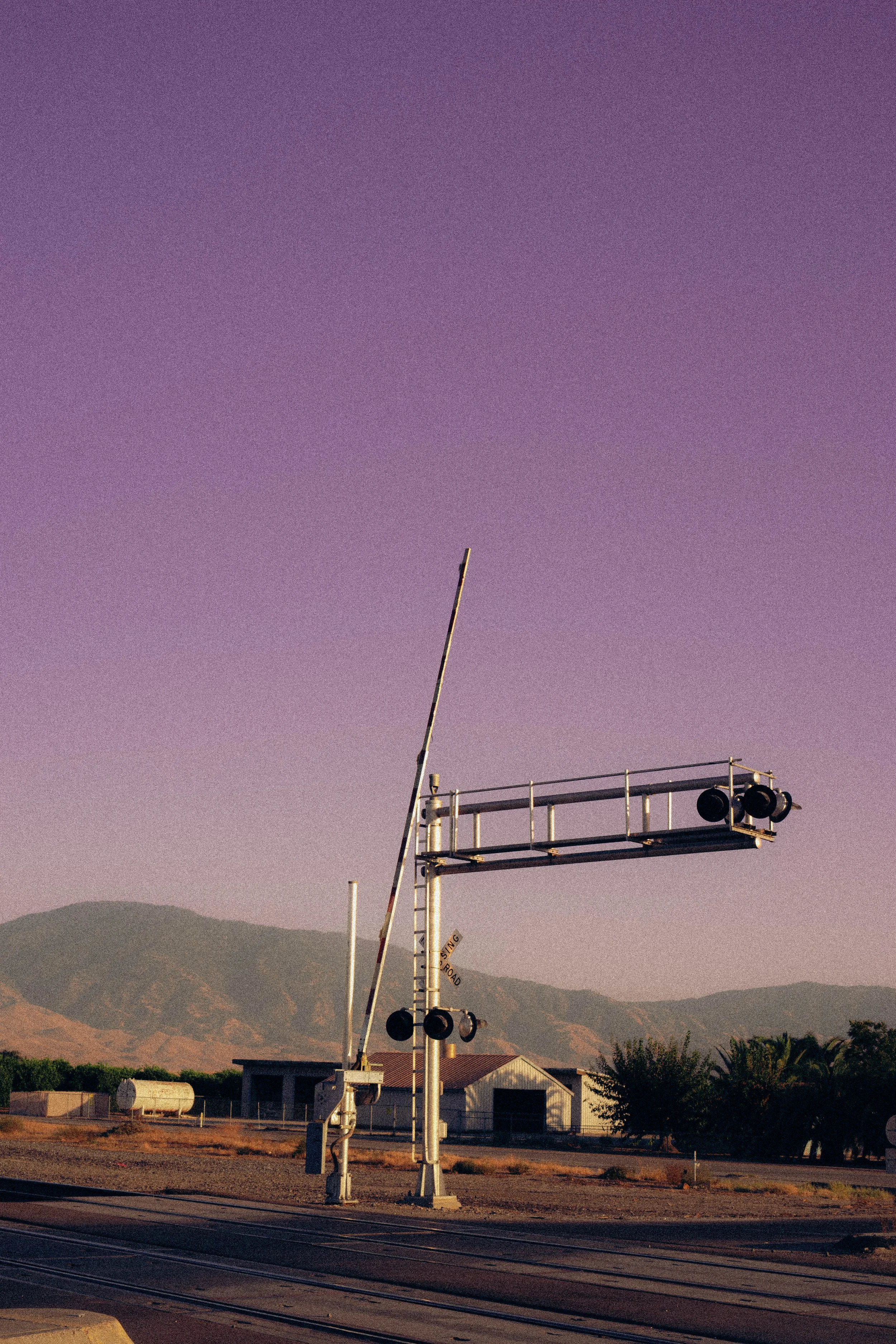 Train crossing gate with a raised barrier arm, signals, and lights, located on railroad tracks with a rural background featuring hills, trees, and a building under a purple-tinged sky.