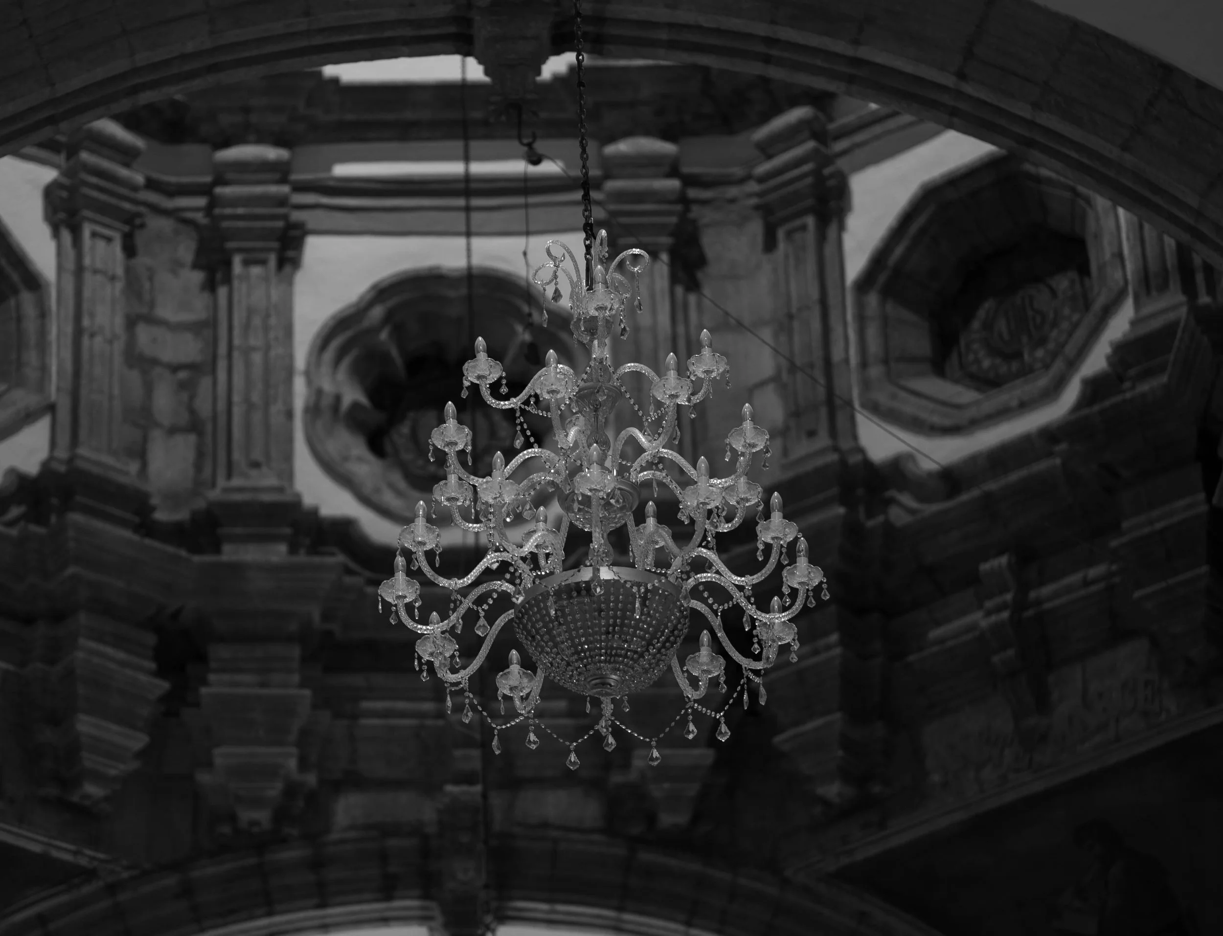 Chandelier hanging from ornate ceiling with architectural details, in black and white.
