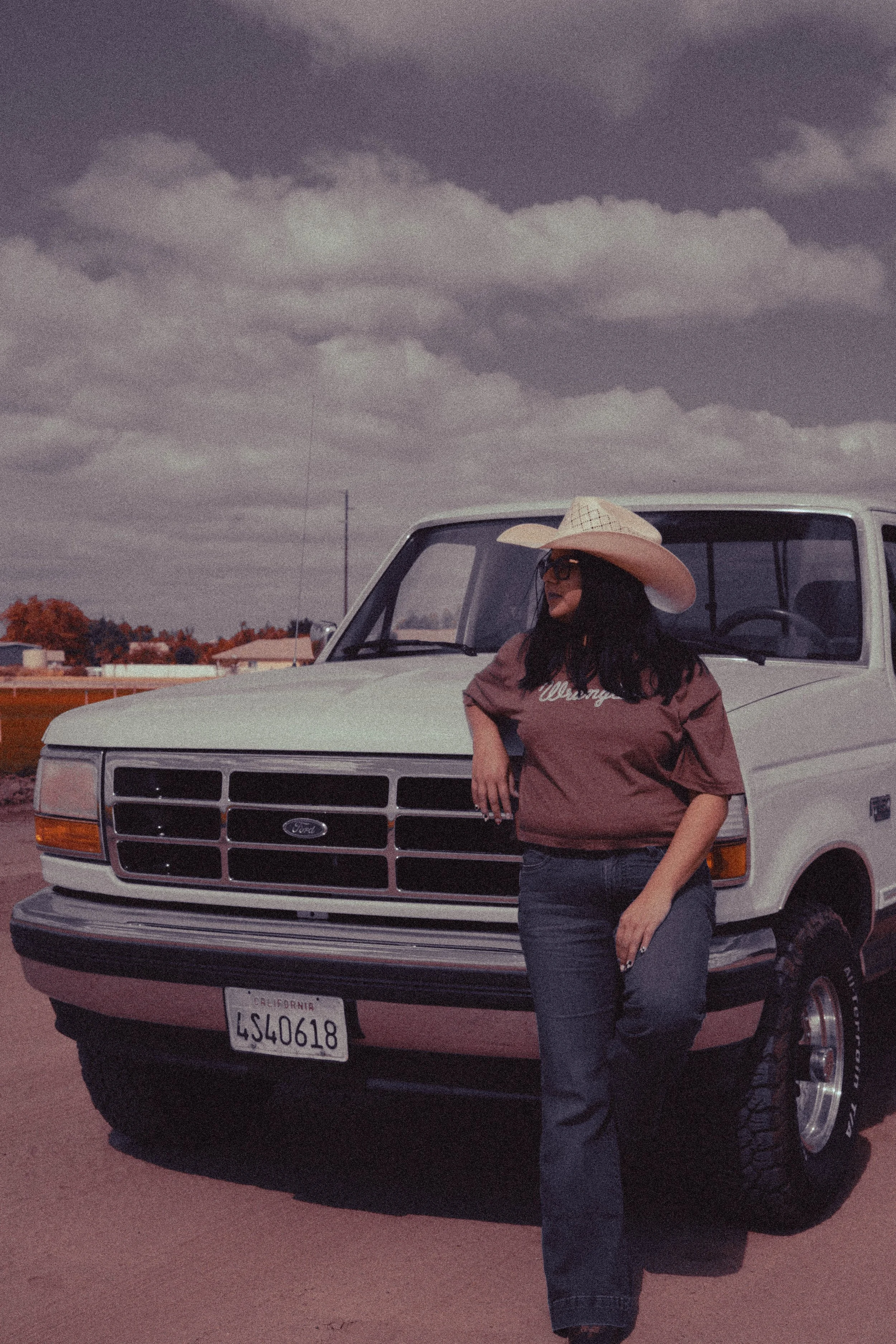 A woman wearing sunglasses, a large sunhat, and a brown t-shirt with 'Wrangler' written on it, leaning against a white Ford truck with a California license plate, under a cloudy sky.