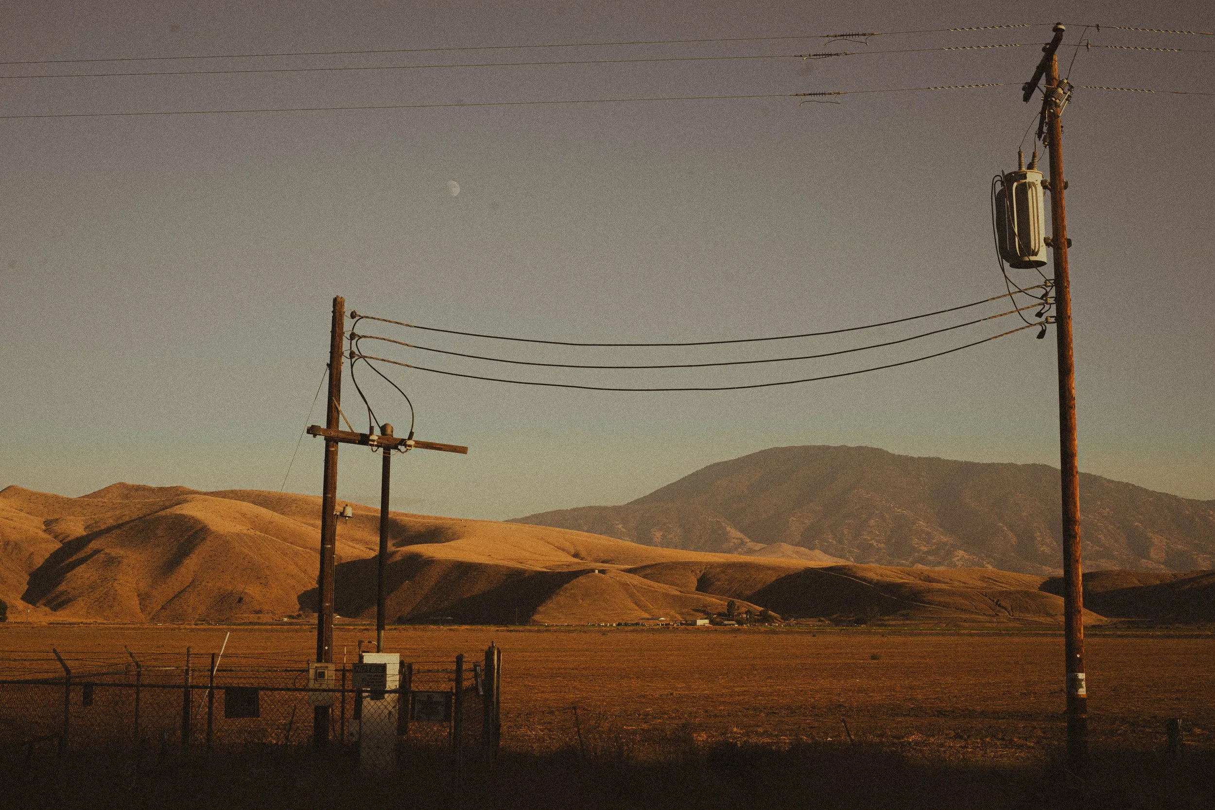 Electric poles in a rural landscape with mountains and a visible moon in the sky.