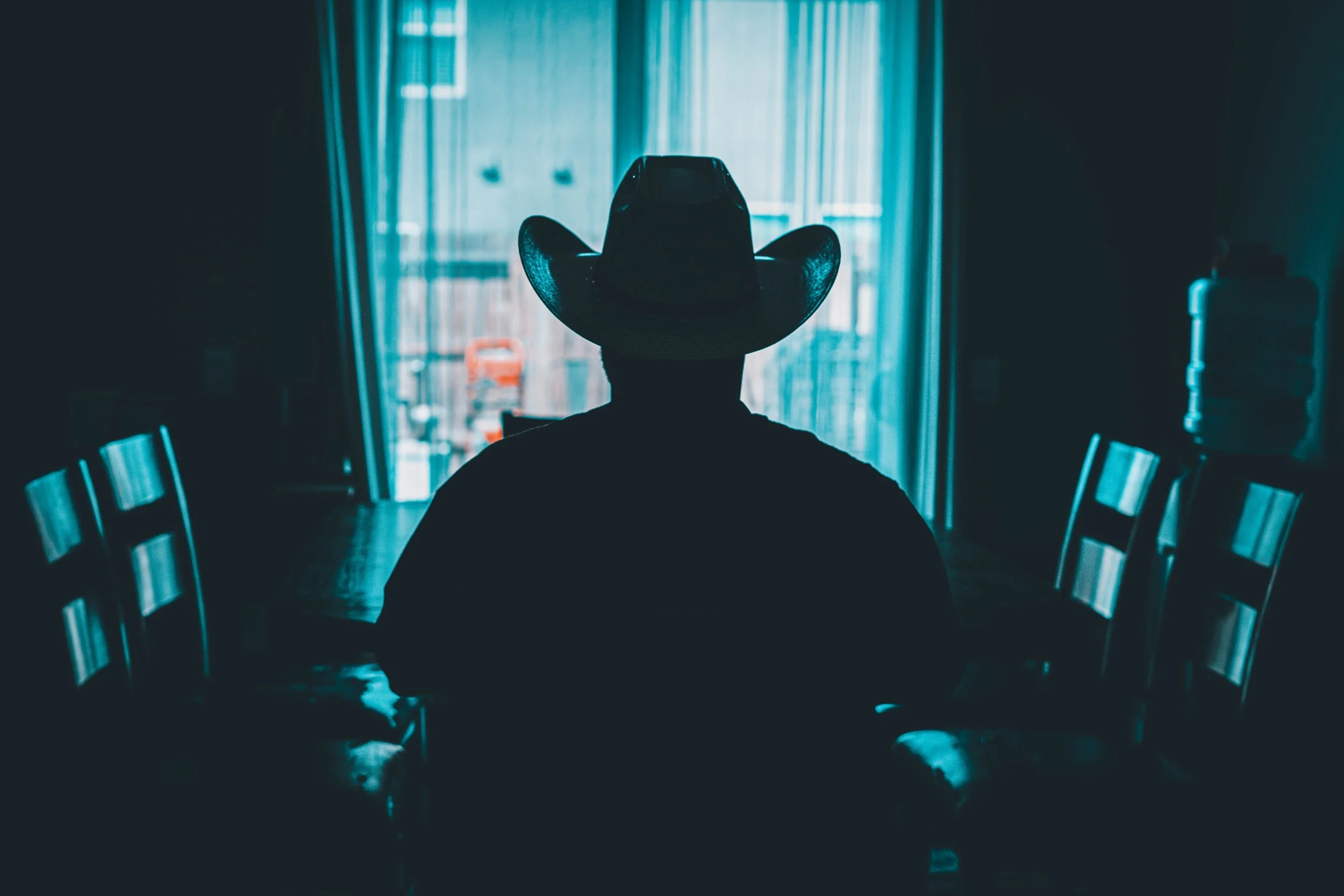 Silhouette of a person wearing a cowboy hat sitting at a dining table inside a dimly lit room with a large window or door in the background.
