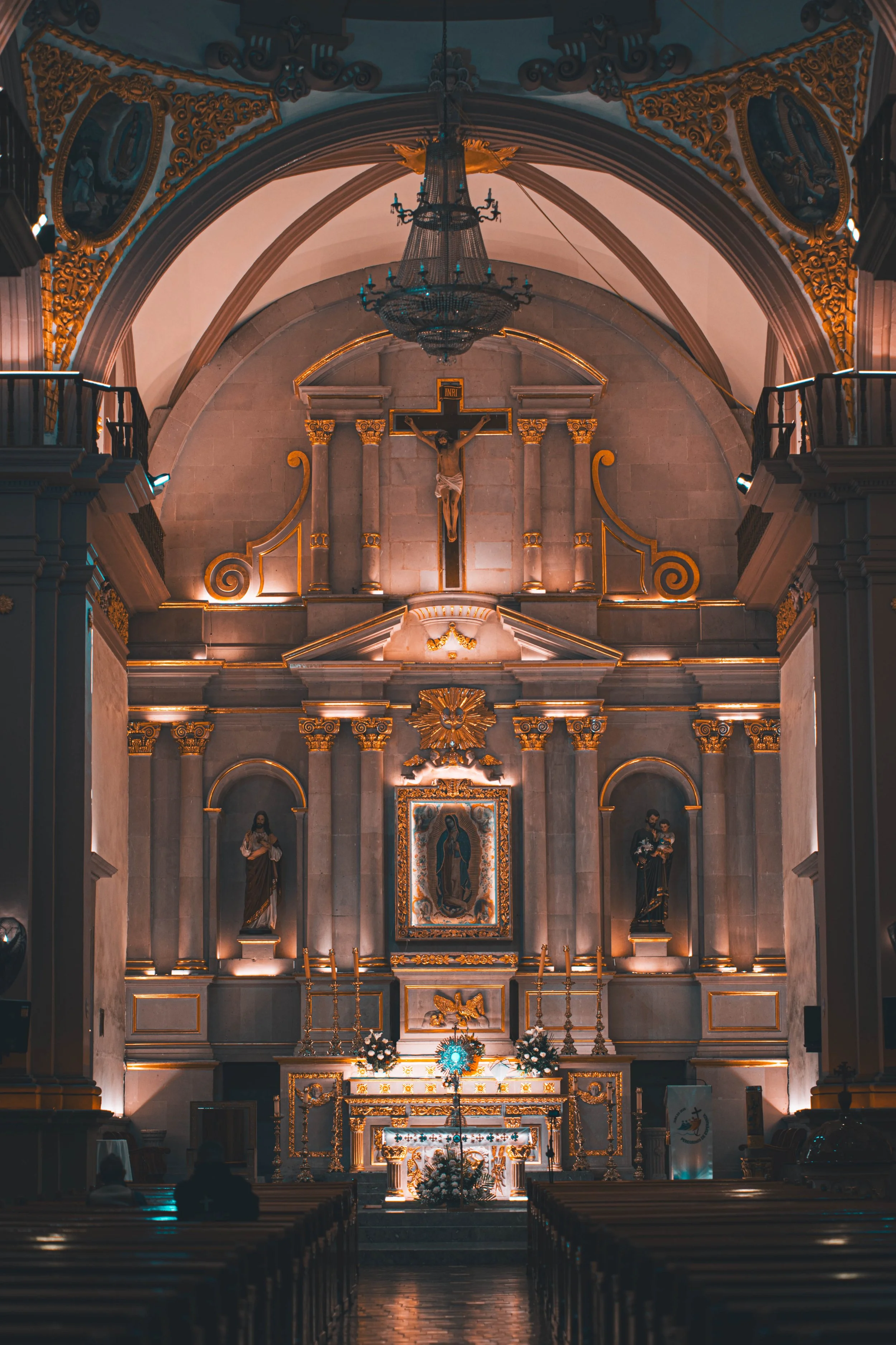 Interior of a church altar with religious statues, gold accents, crucifix, and soft lighting.