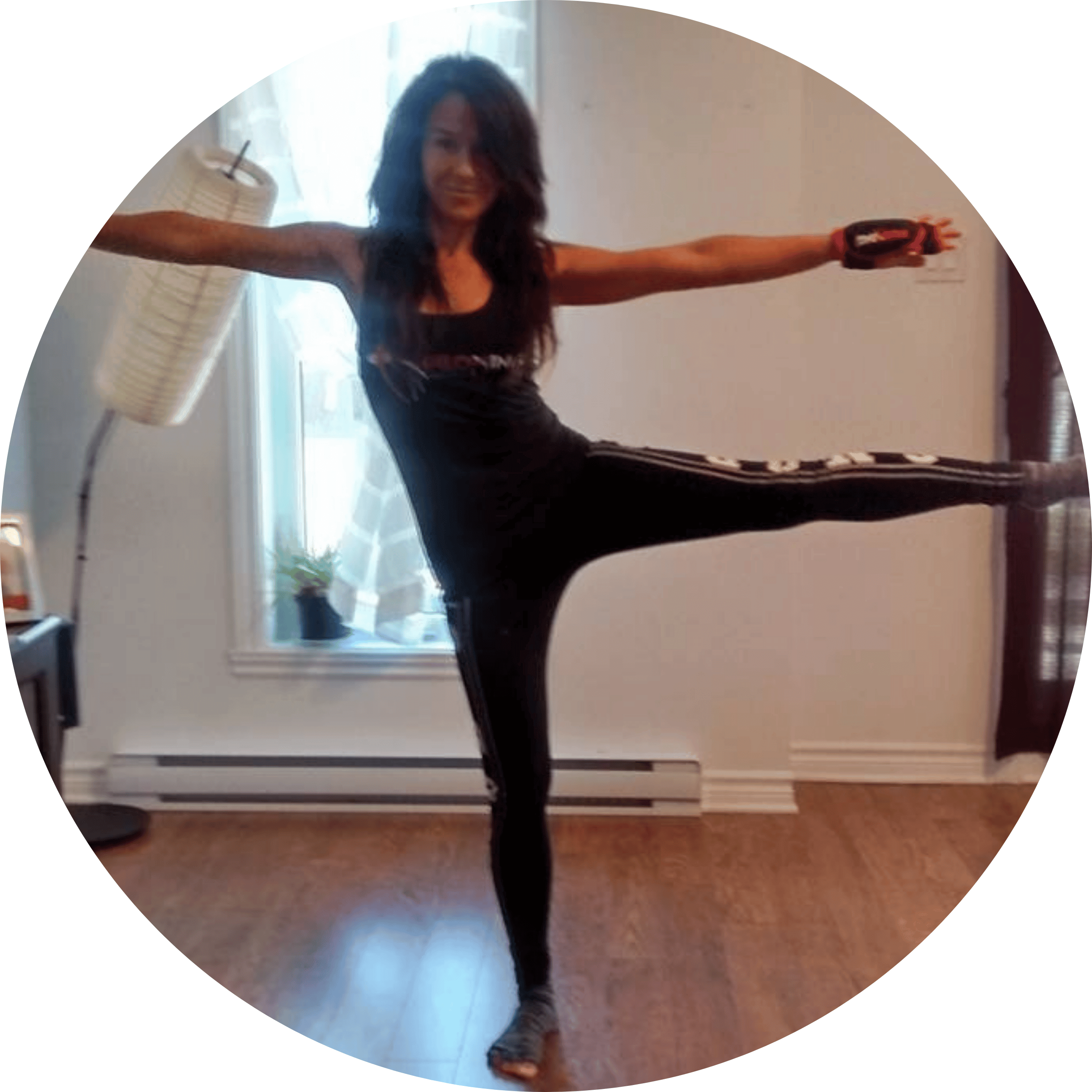 Woman in black athletic clothing standing on one leg with arms outstretched, balancing indoors near a window.