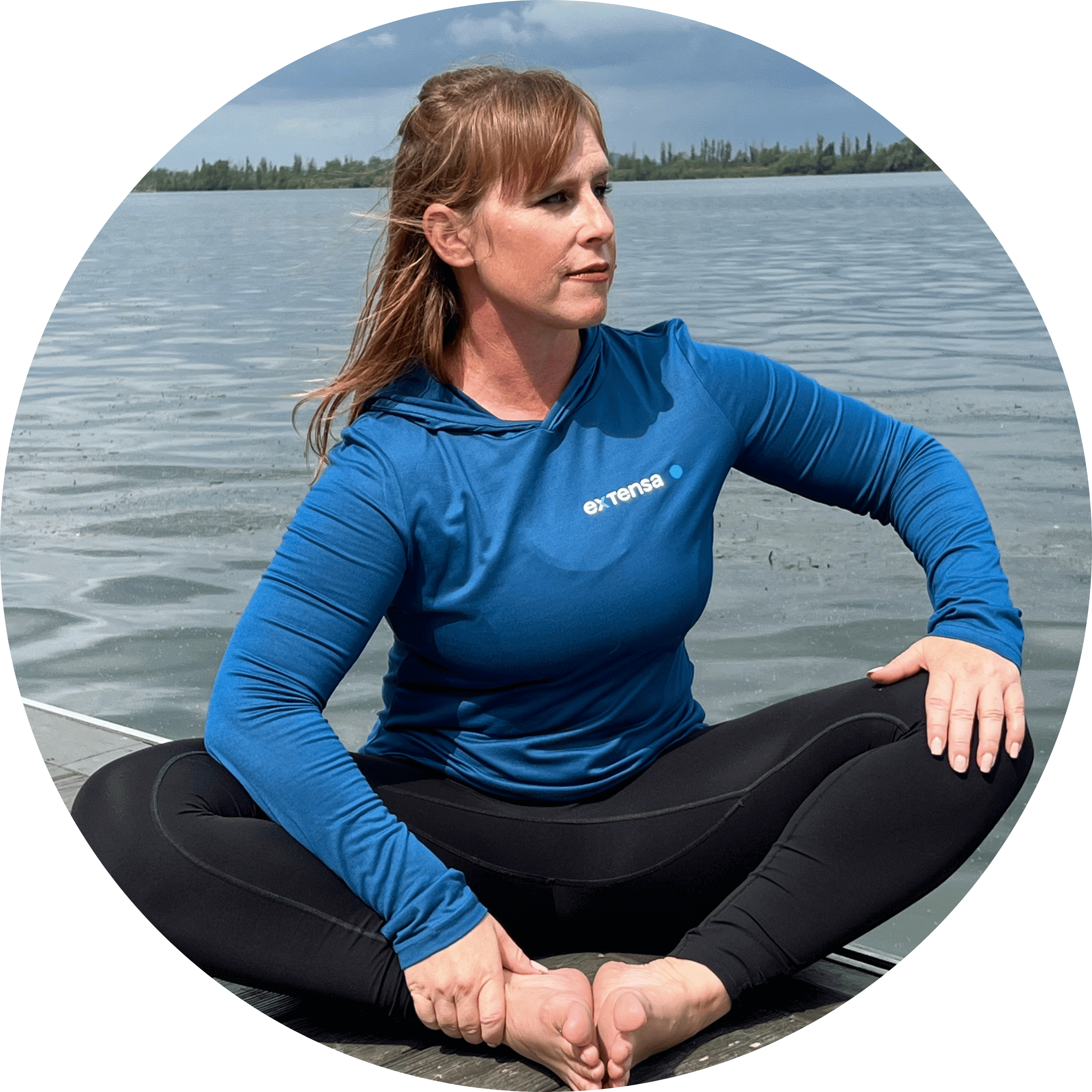 A woman with red hair practicing yoga on a dock near a body of water, wearing a blue long-sleeve shirt with a logo and black leggings.