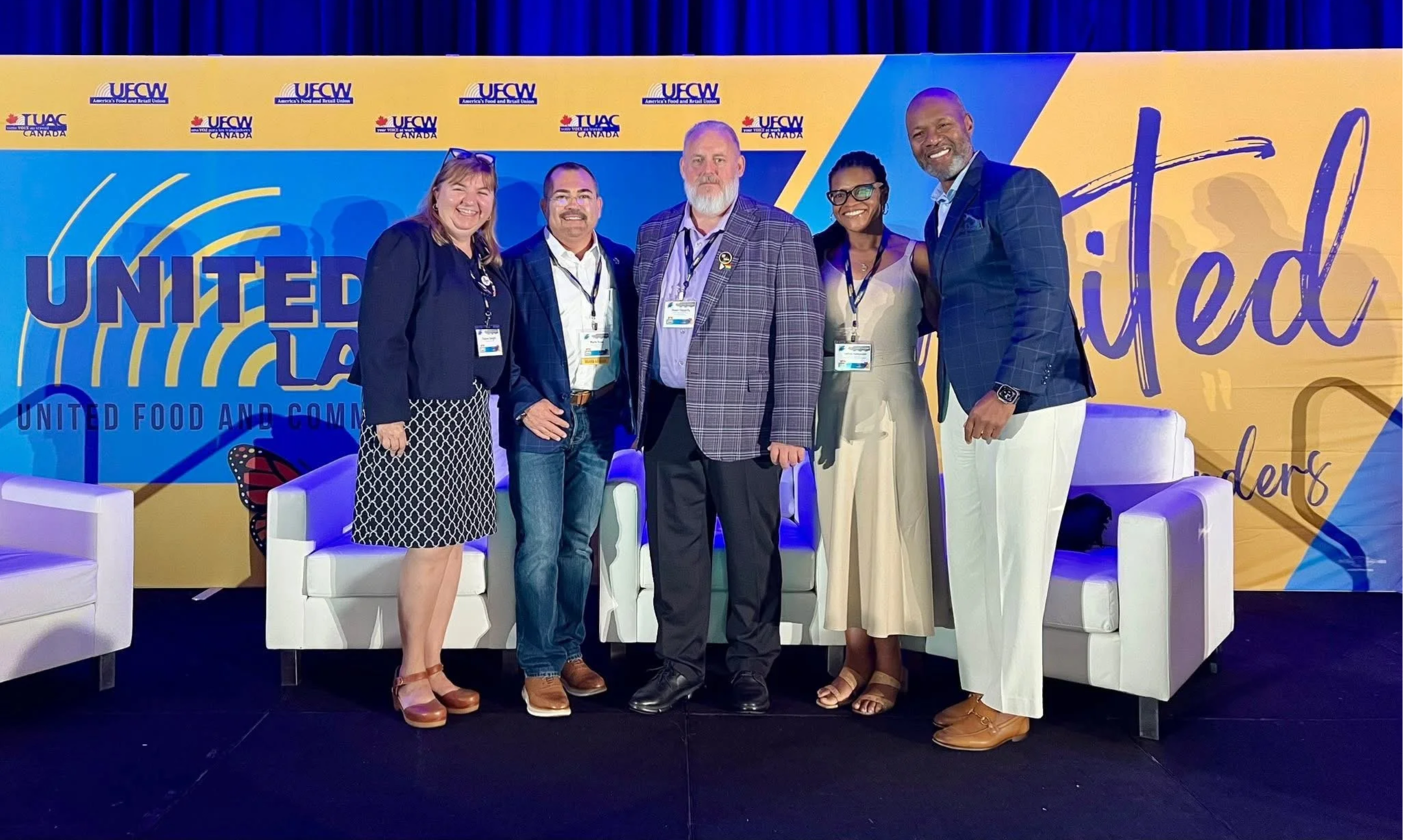 Five diverse individuals standing on stage in front of a blue and yellow UNited themed backdrop, smiling for a photo at a conference or event.