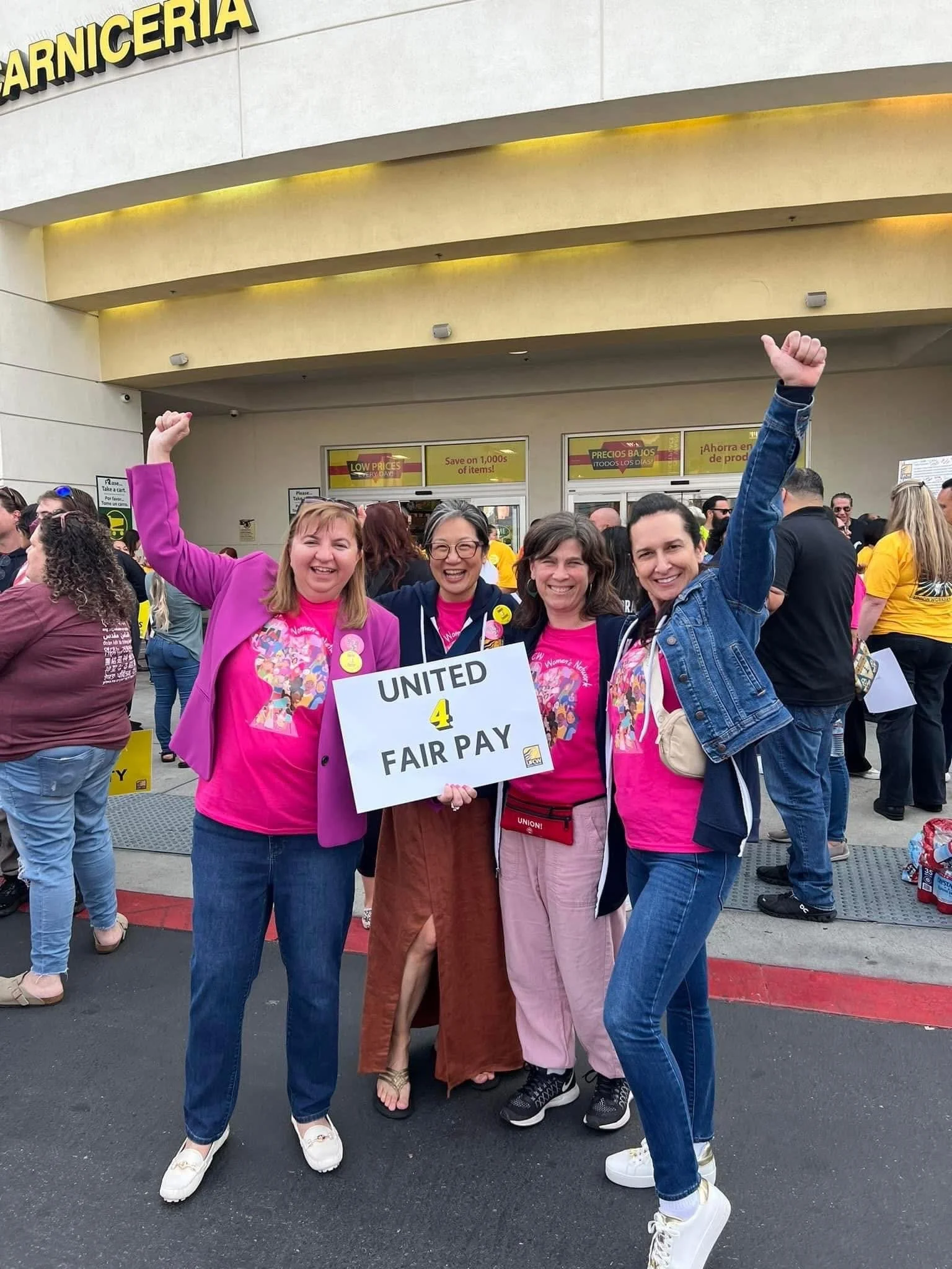 Four women standing outside a store in a protest, holding a sign that reads "UNITED 4 FAIR PAY," wearing pink shirts, with one woman raising her fist in the air.