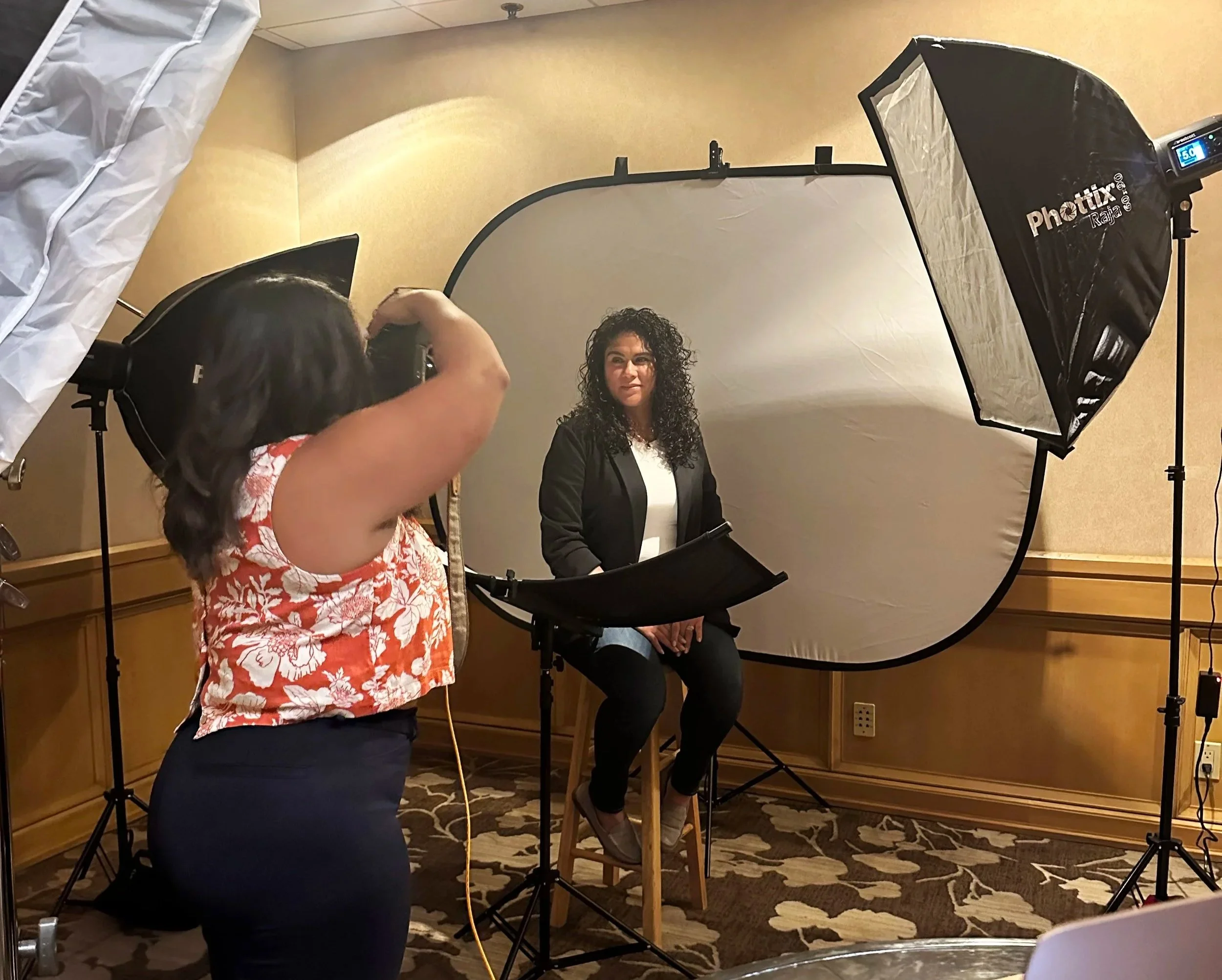 A woman with curly hair sitting in front of a photography backdrop, being photographed by a photographer in a studio setting with lighting equipment.