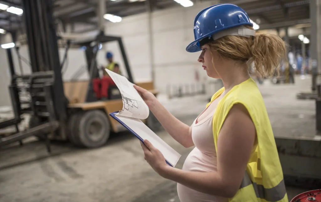 Pregnant woman in a blue hard hat and yellow safety vest holding and reviewing a blueprint inside a warehouse or manufacturing facility with forklifts and workers in the background.