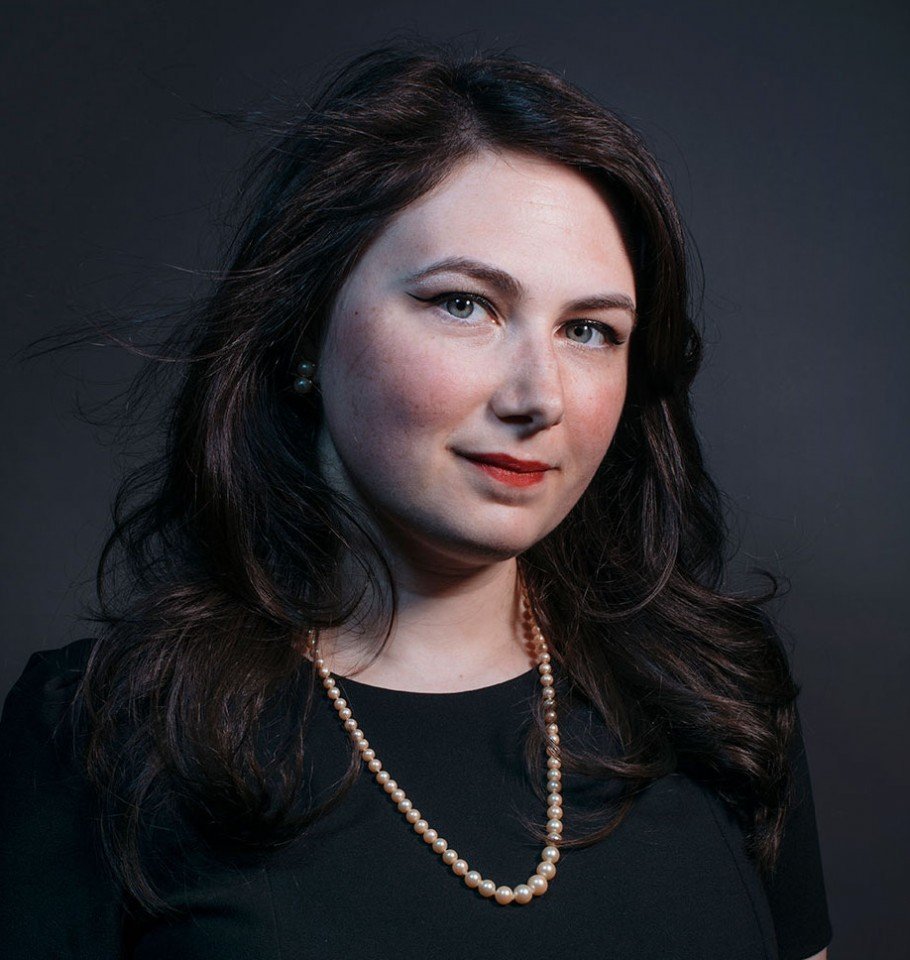 Portrait of a woman with dark brown hair, light skin, wearing pearl earrings and a pearl necklace, dressed in a black top, against a dark background.
