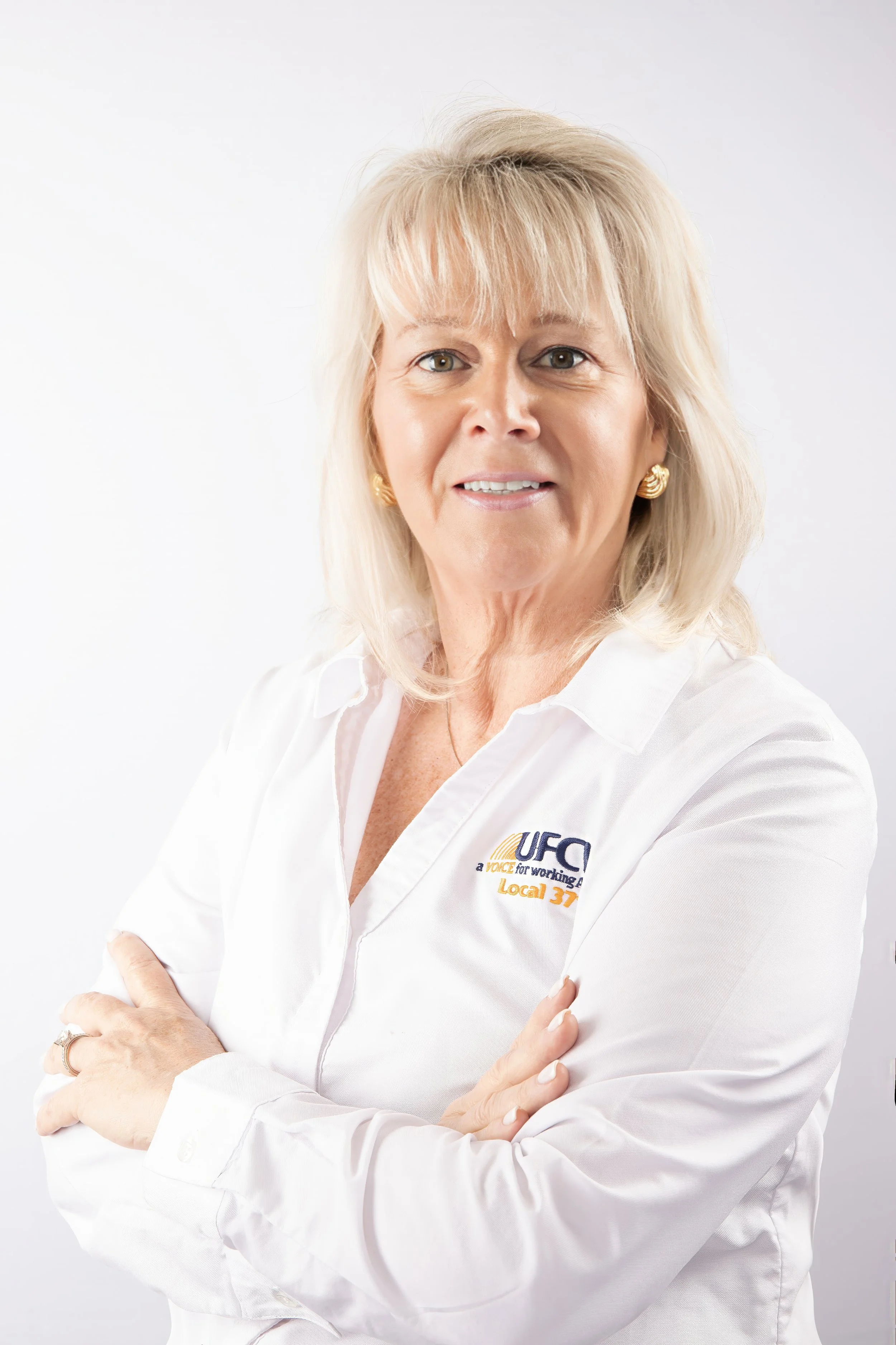 A woman with blonde hair, wearing a white shirt with a logo, standing against a white background with her arms crossed, smiling at the camera.