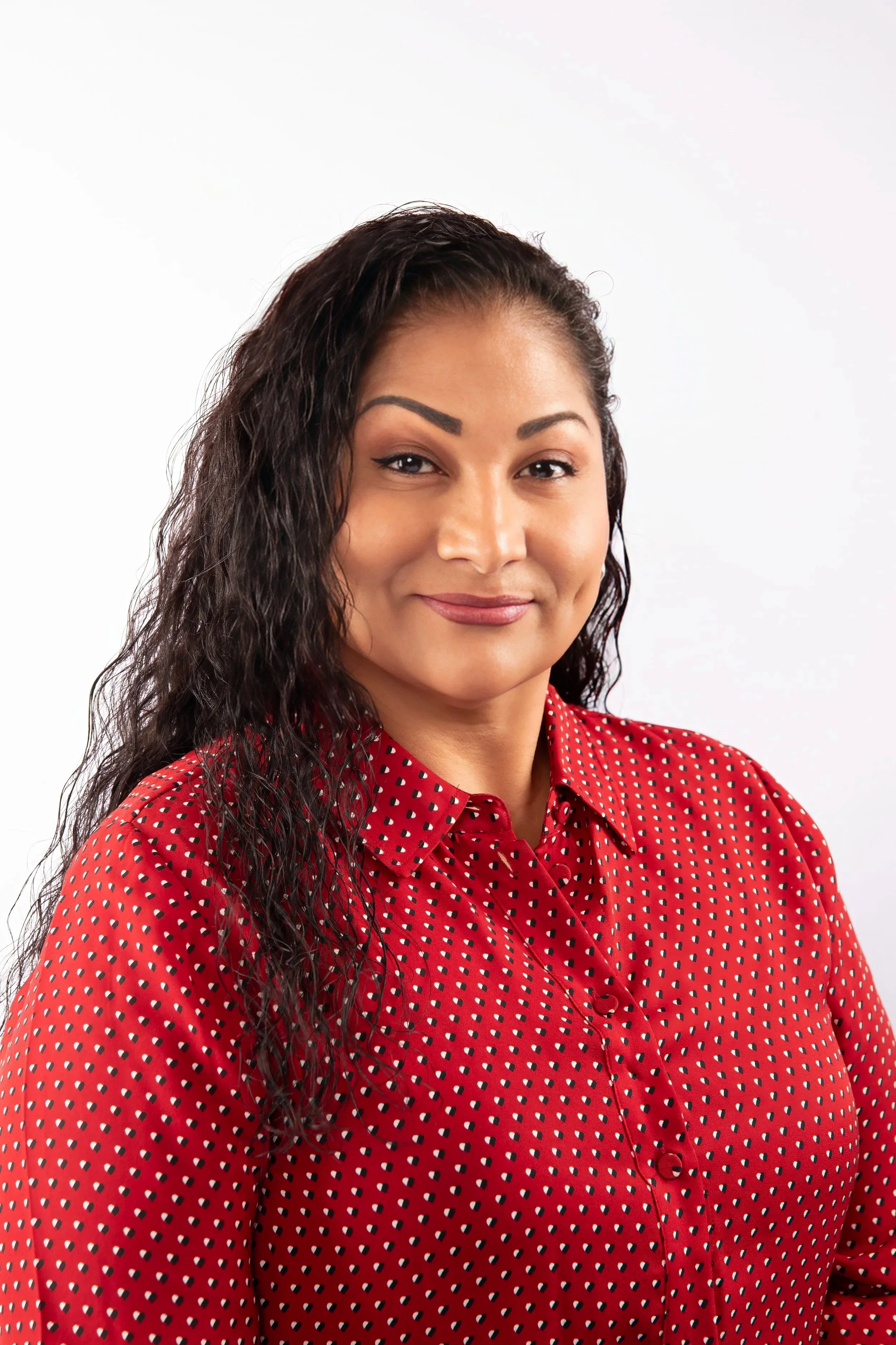 A woman with dark, wavy hair wearing a red shirt with white polka dots, smiling softly against a plain white background.