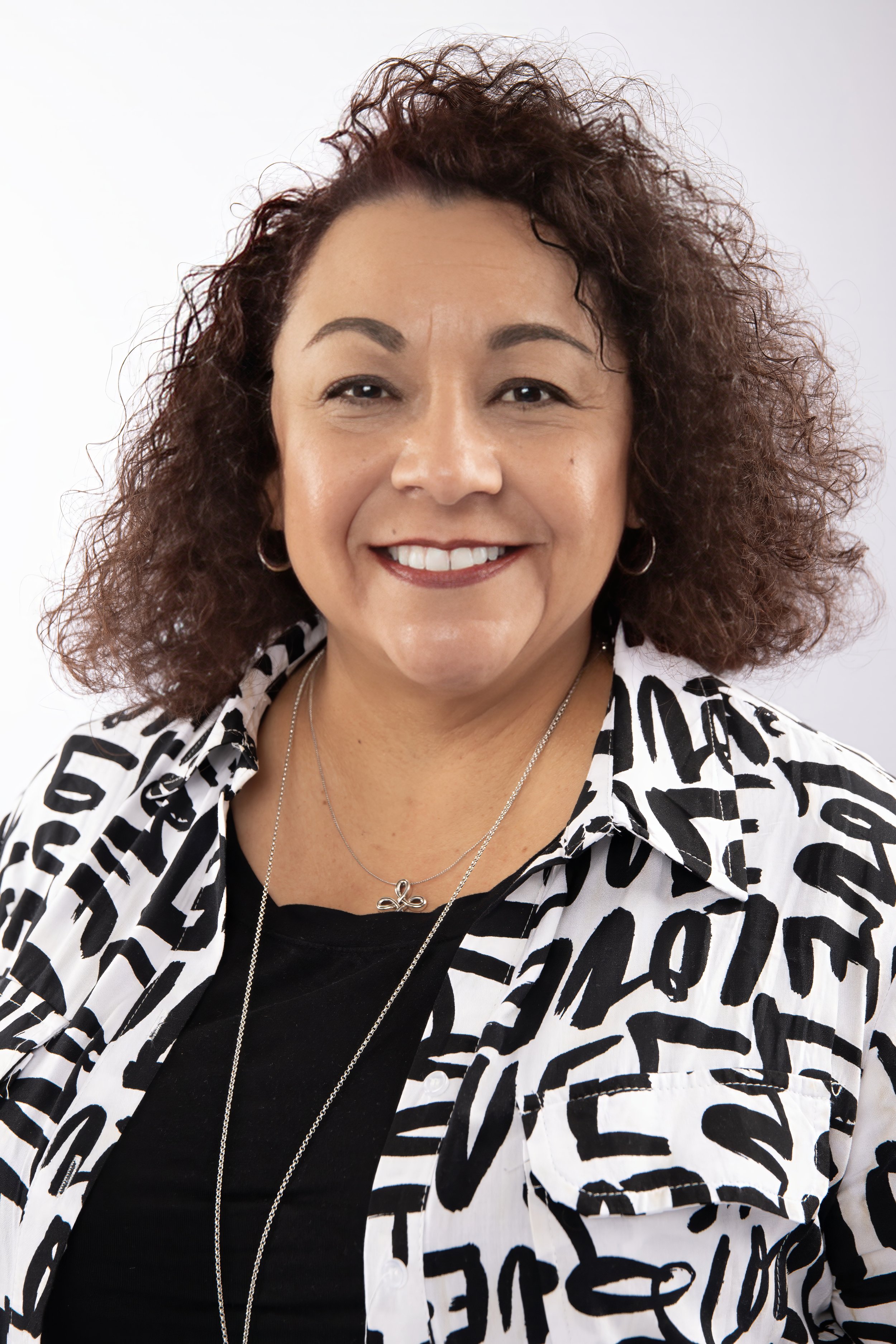 A smiling woman with curly brown hair, wearing a black top, a white jacket with black abstract lettering, and layered jewelry, standing against a plain white background.