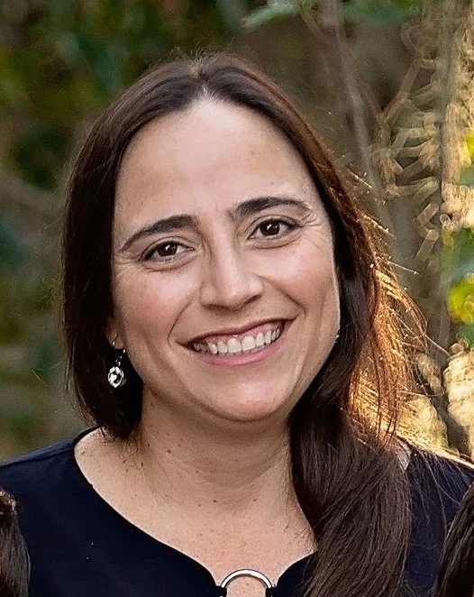 Close-up of a woman with brown hair, smiling, wearing earrings and a black top, outdoors with blurred greenery in the background.